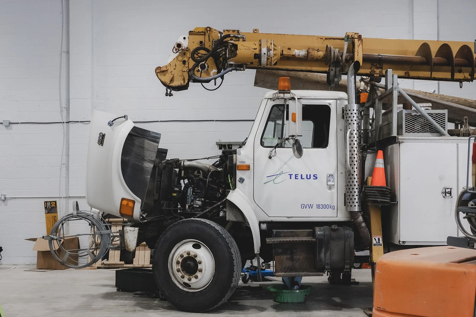 A TELUS utility truck inside a warehouse with its hood open and various equipment, including a yellow crane arm, placed on the truck and nearby.