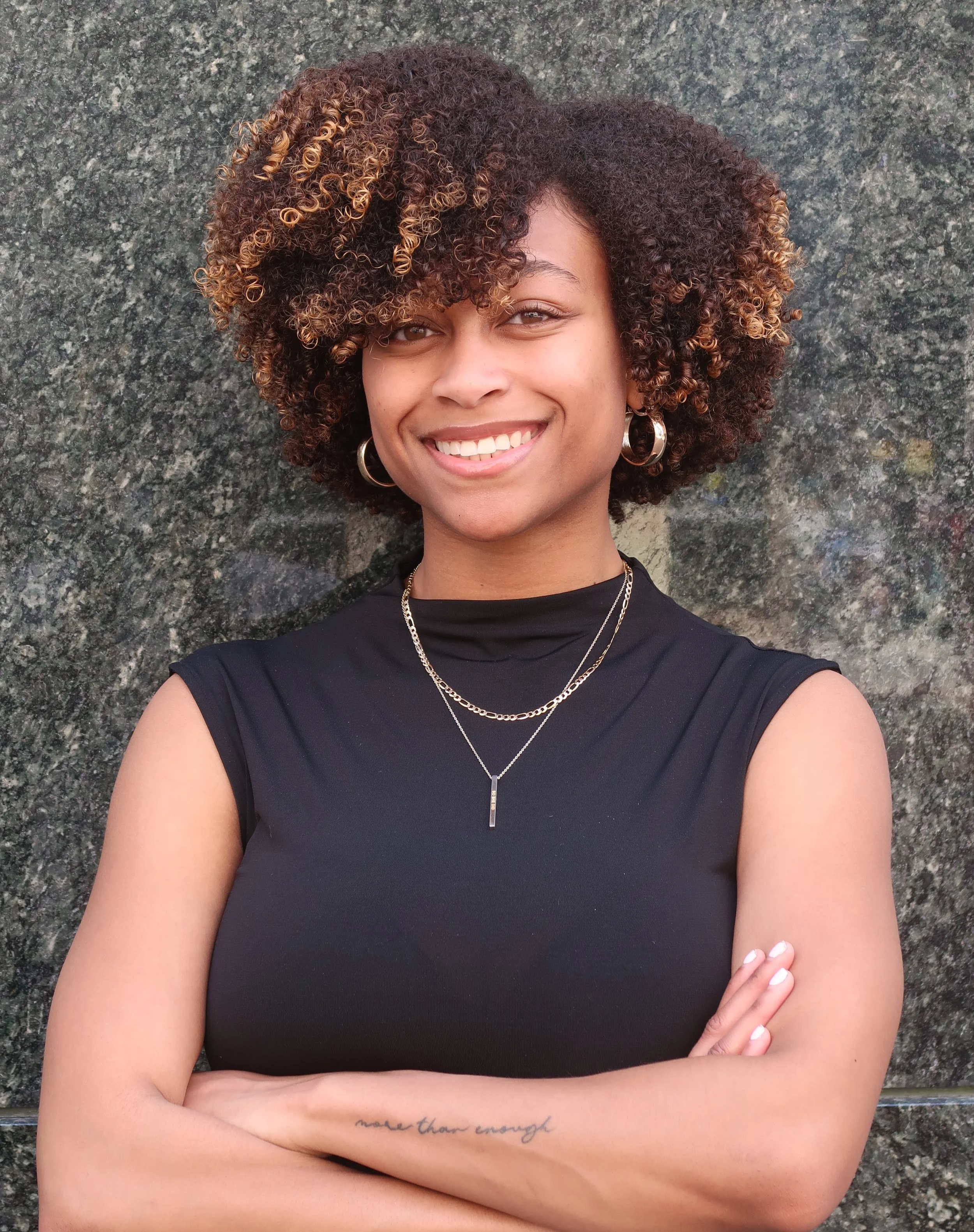 A woman with a smile, cross arms, wearing a black sleeveless top, gold hoops, layered necklaces, and a tattoo on her right forearm, standing against a dark stone background.