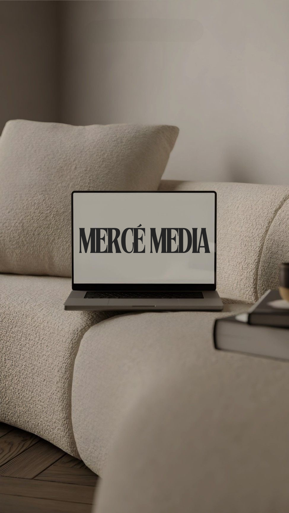 A laptop on a beige textured sofa displays the text 'MERCE MEDIA' on its screen, with a beige cushion and a bookshelf or table with books nearby.