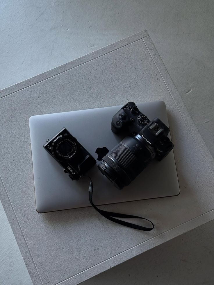 A silver laptop with a Canon camera, a camera lens, and a strap placed on top of it, all resting on a white textured surface.