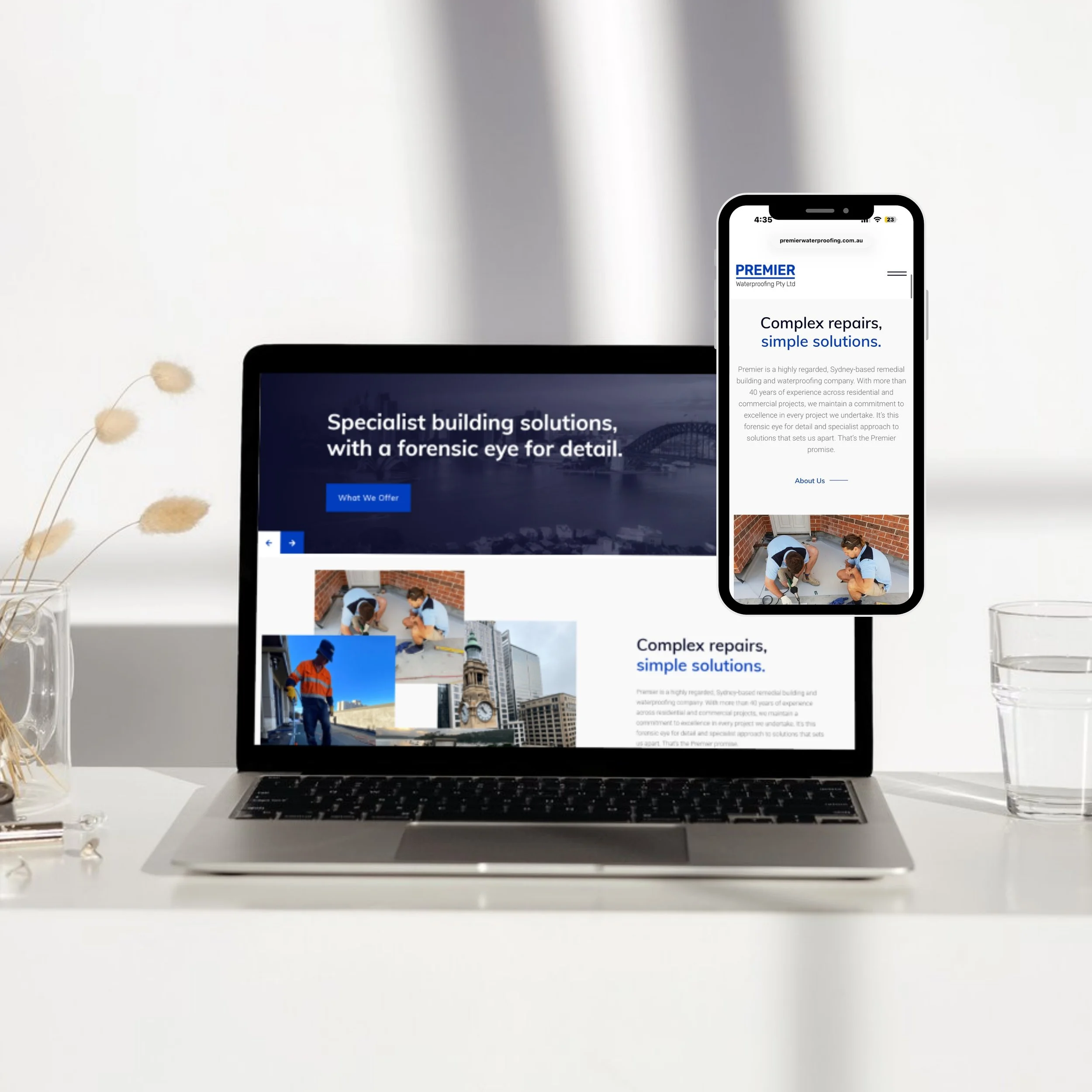 Laptop and smartphone displaying a website about building solutions and repairs, with images of workers and cityscapes, on a white desk with a glass of water and a vase with dried plants.