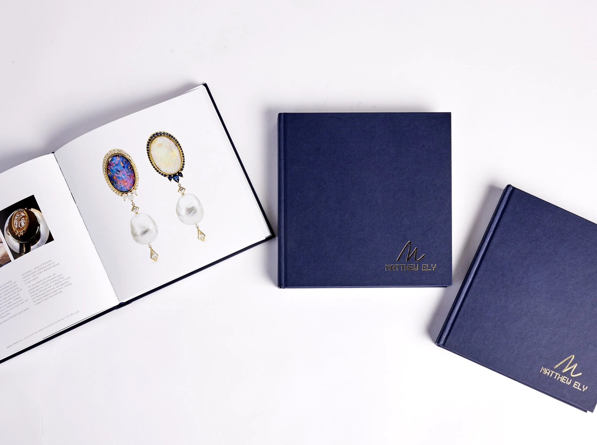 Open jewelry catalog displaying a pair of colorful earrings with pearl drops, surrounded by two closed blue books with 'Matthew Ely' written in gold on the covers, set on a white surface.