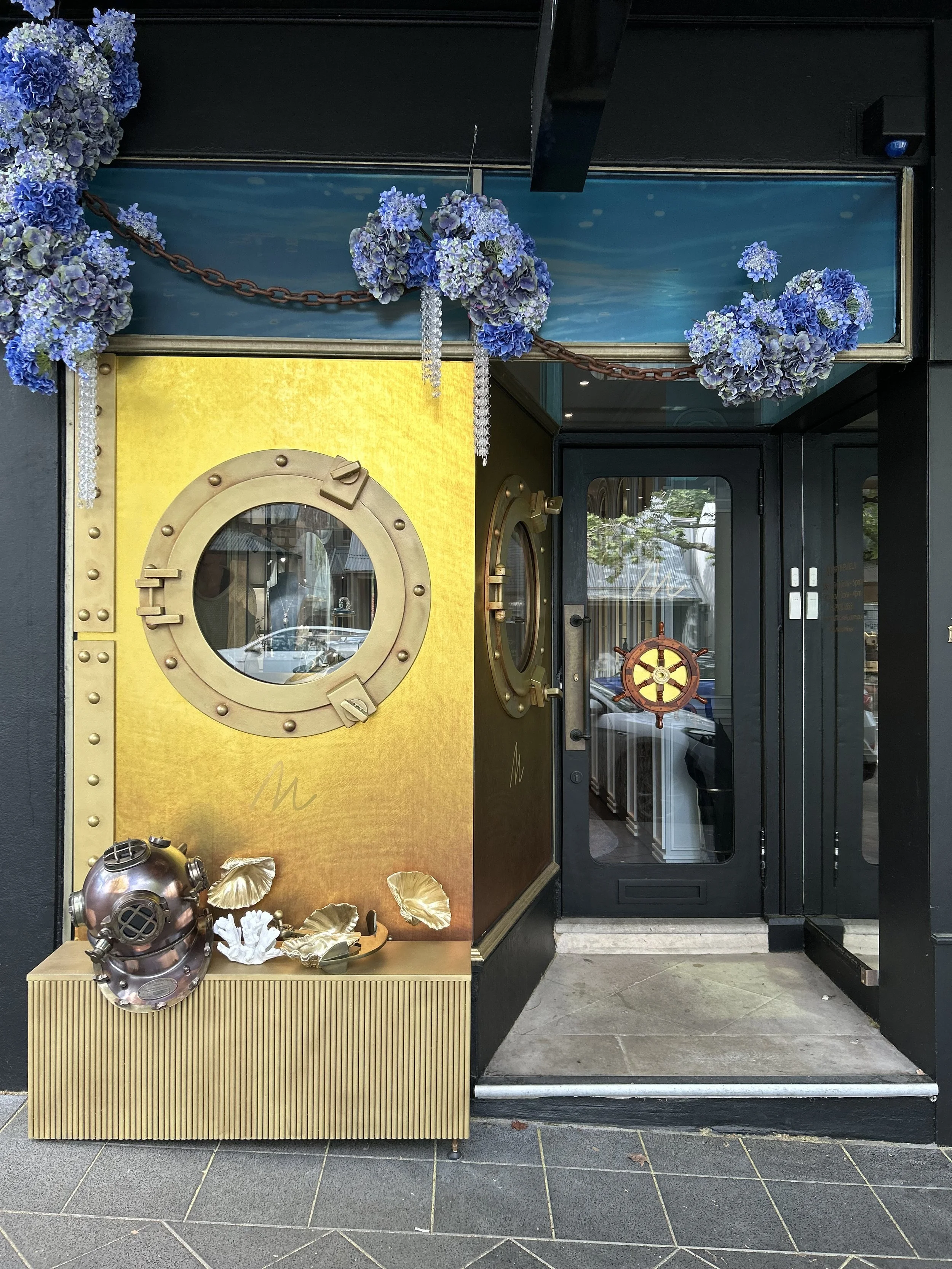 Storefront decorated with blue hydrangea flowers, gold accents, and nautical-themed decor, including a porthole window and a ship's wheel on the door.
