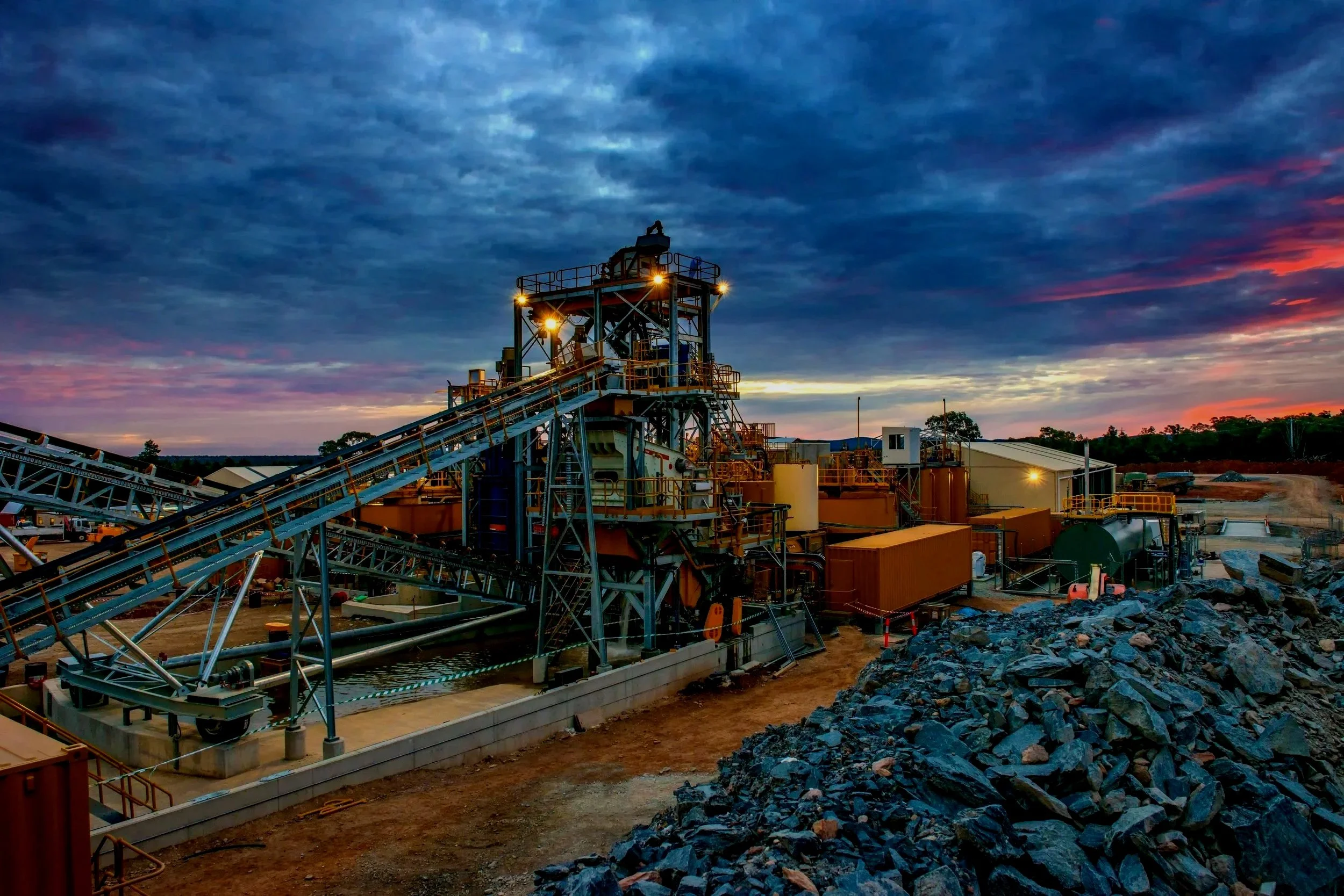 An industrial mining site with conveyor belts, processing equipment, and piles of rocks under a cloudy sunset sky.
