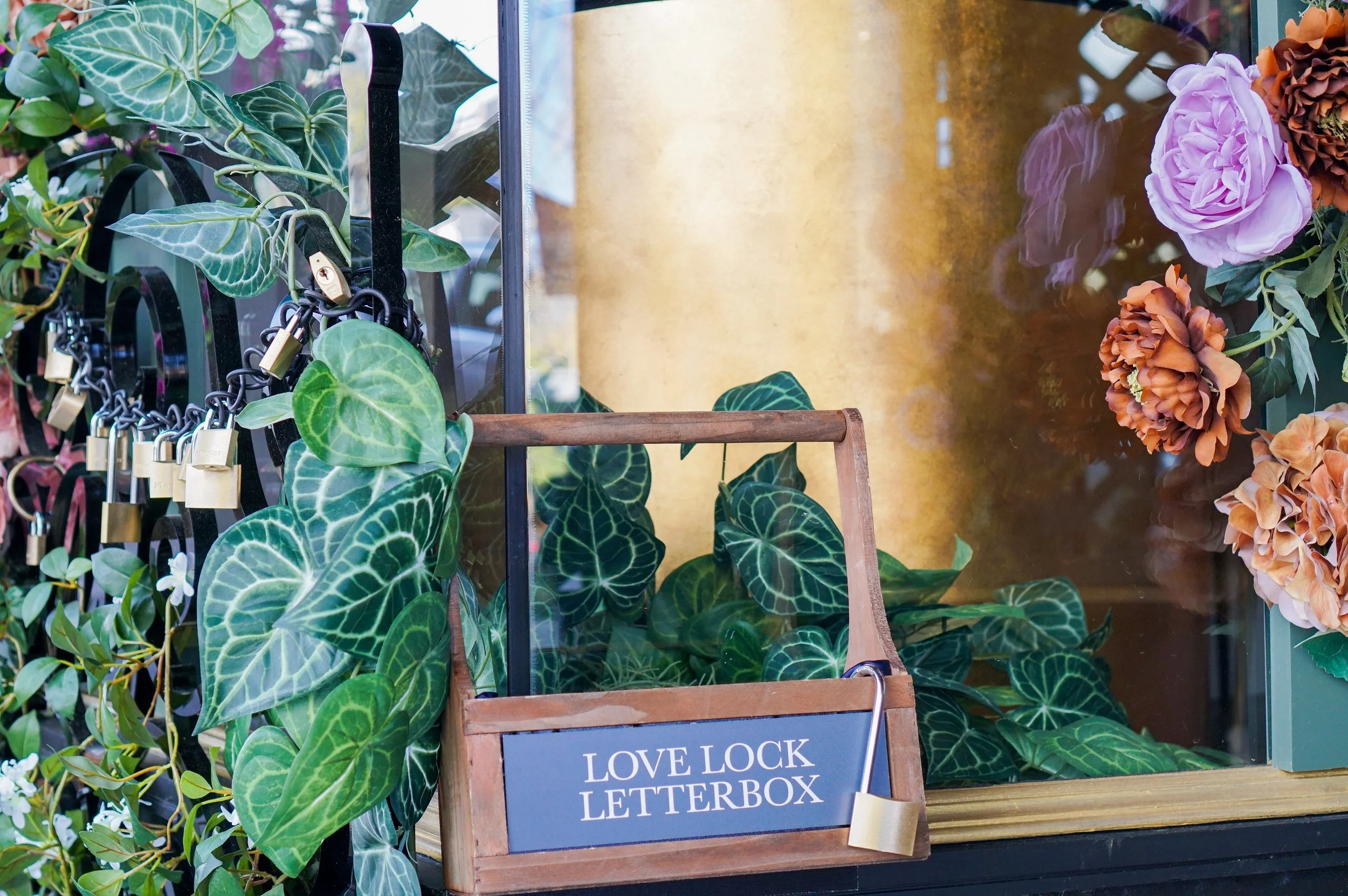 A storefront display with green leafy plants, a wooden love lock letterbox with a padlock attached, and pink and brown flowers on the right.