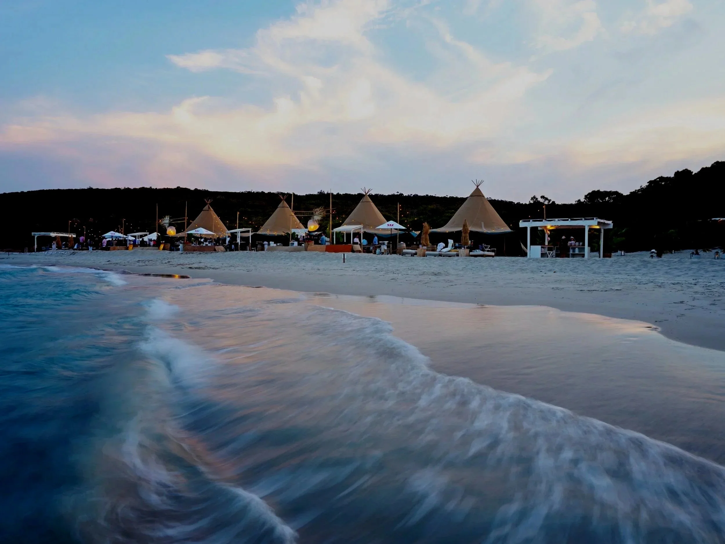 Beachside bar with multiple sand-colored tents, umbrellas, and seating areas at dusk or sunset, overlooking the ocean waves and a hill in the background.