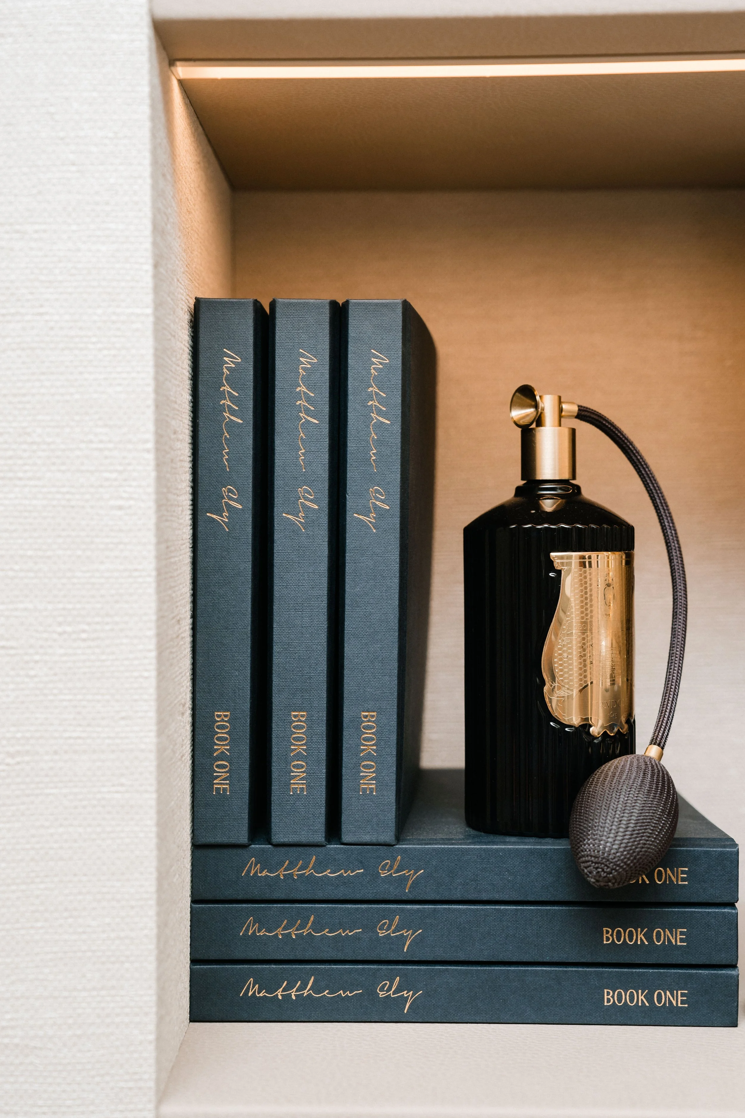 Three dark blue hardcover books titled 'Matthew Ely' and 'Book One' stacked upright, a black and gold perfume atomizer, and a sound sponge on top of the books.