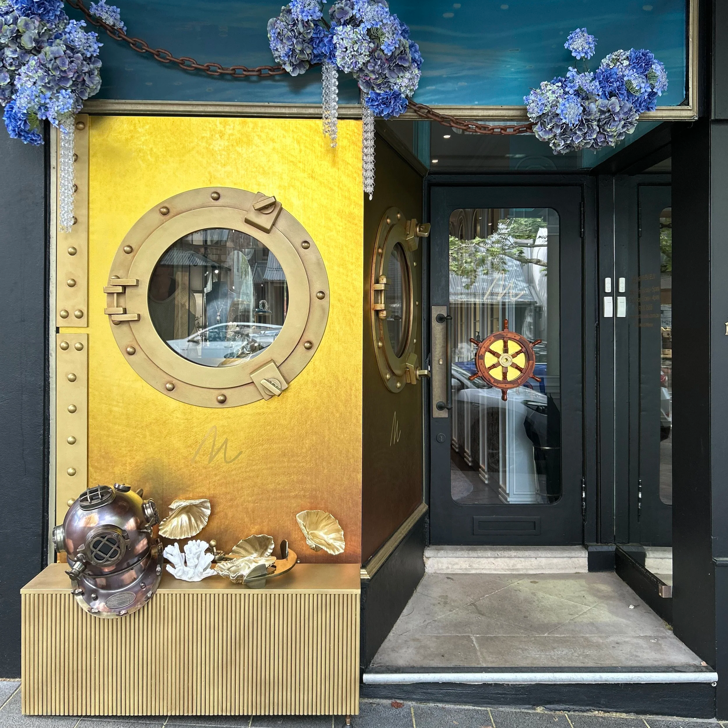 Decorative entrance door with a maritime theme, featuring a porthole window, a ship's steering wheel on the glass door, and nautical decorations including a vintage diving helmet, gold-colored seashell sculptures, and flowers with blue hydrangeas at the top.