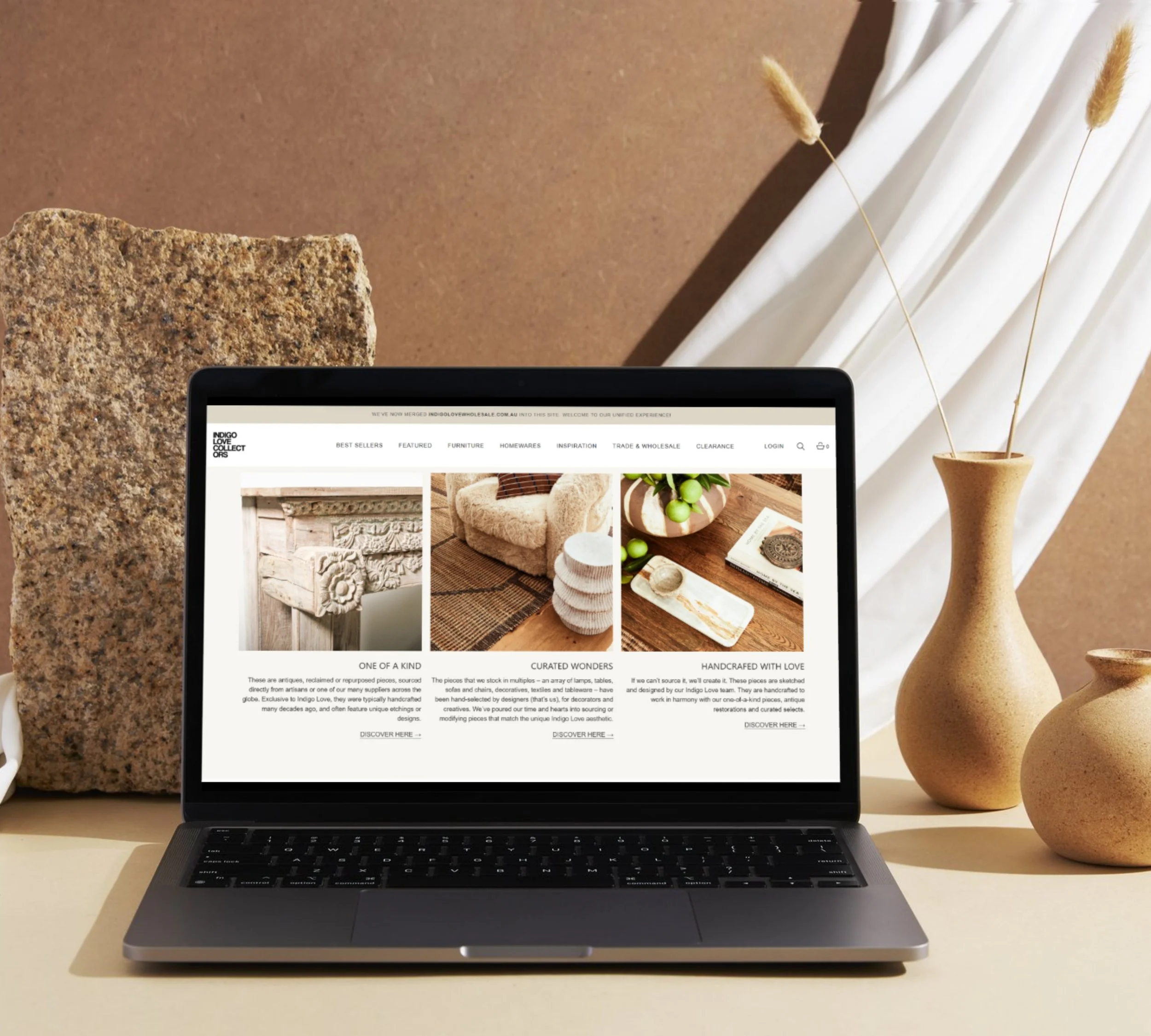 Laptop displaying a website about handmade home decor, placed on a beige surface with decorative vases and large textured stone piece in the background.