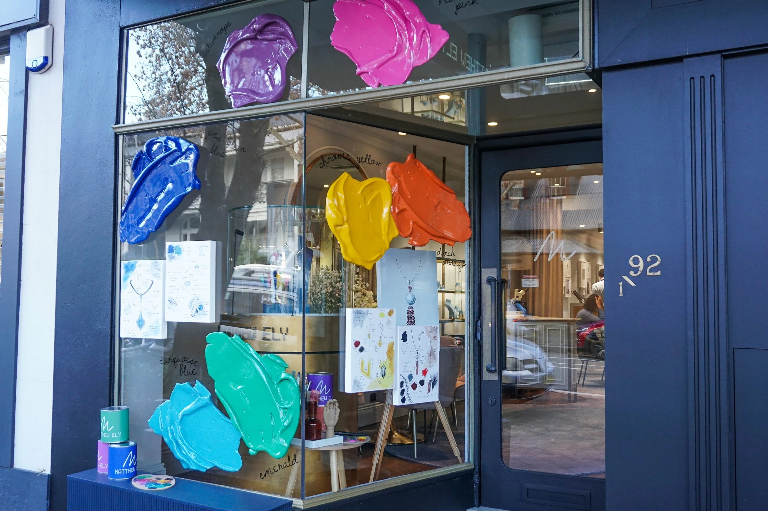 Colorful paint blobs on window display of a jewelry store with necklaces and jewelry sketches inside.