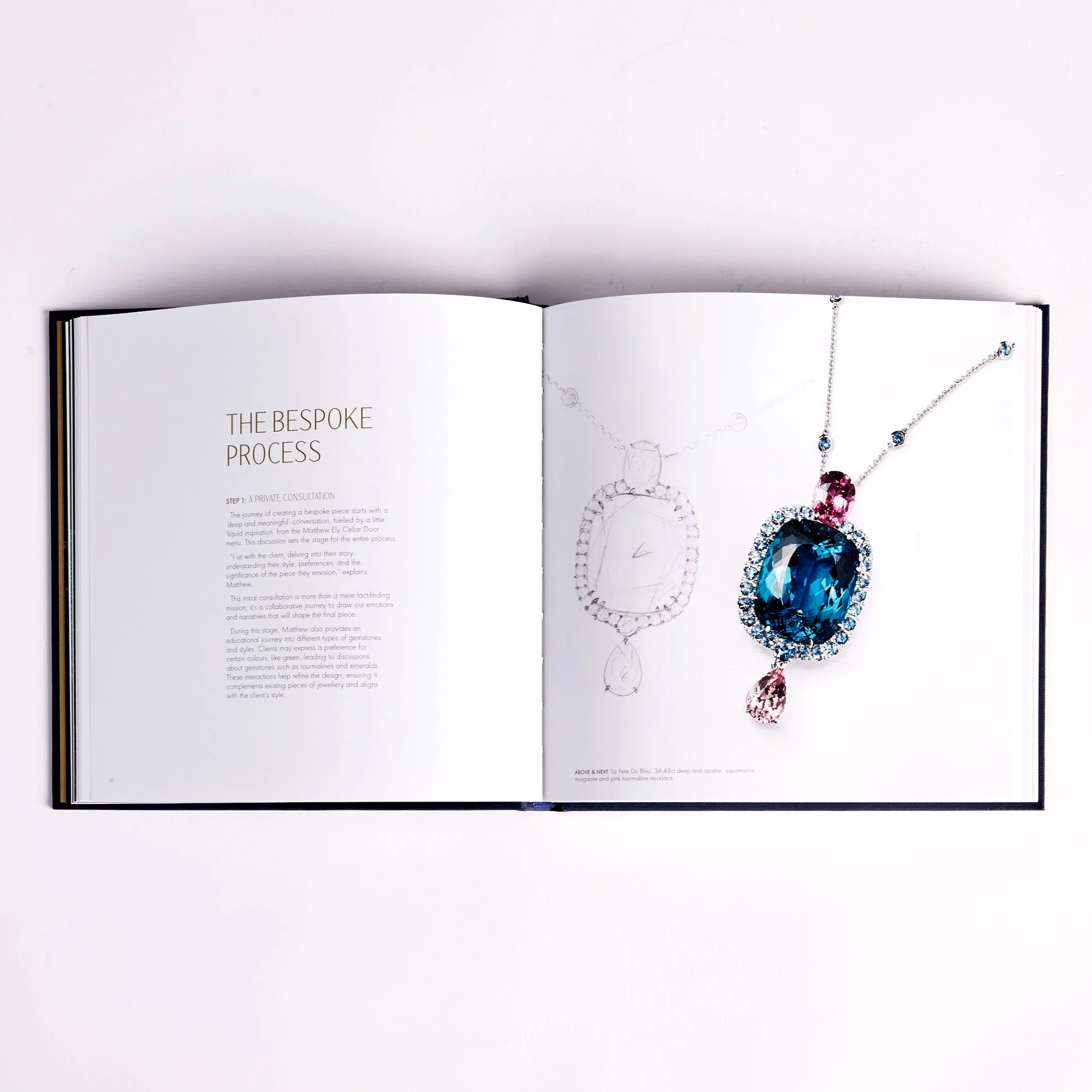 Open jewelry catalog displaying a detailed drawing and a photo of a gemstone necklace with a large blue gemstone surrounded by smaller diamonds and pink gemstones, laid out on white pages.