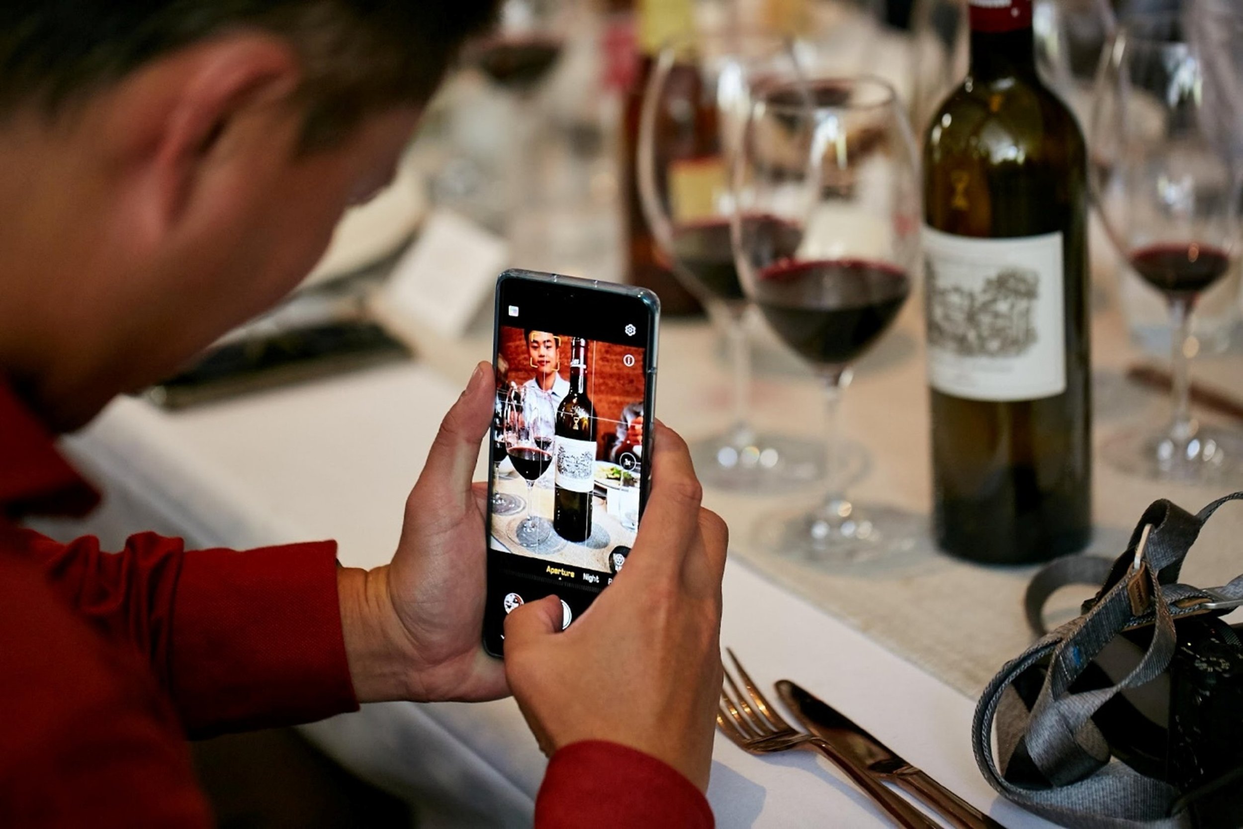 Person taking a photo of a wine bottle and glasses of red wine on a dining table with a smartphone