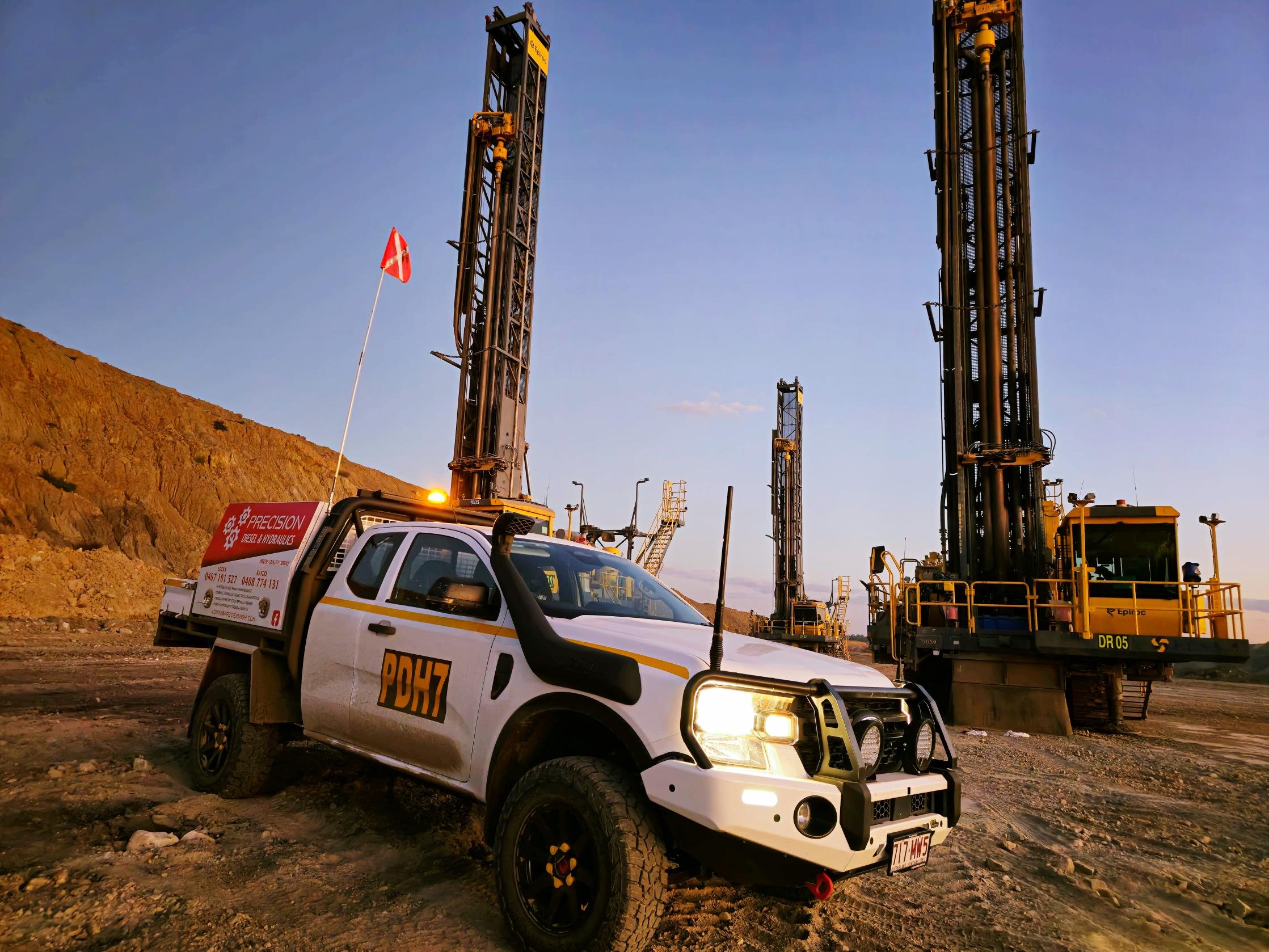 A white utility pickup truck with yellow and black markings and the word 'POHZ' on the side, parked on a dirt road at an industrial site with large machinery and drilling rigs under a clear sky.