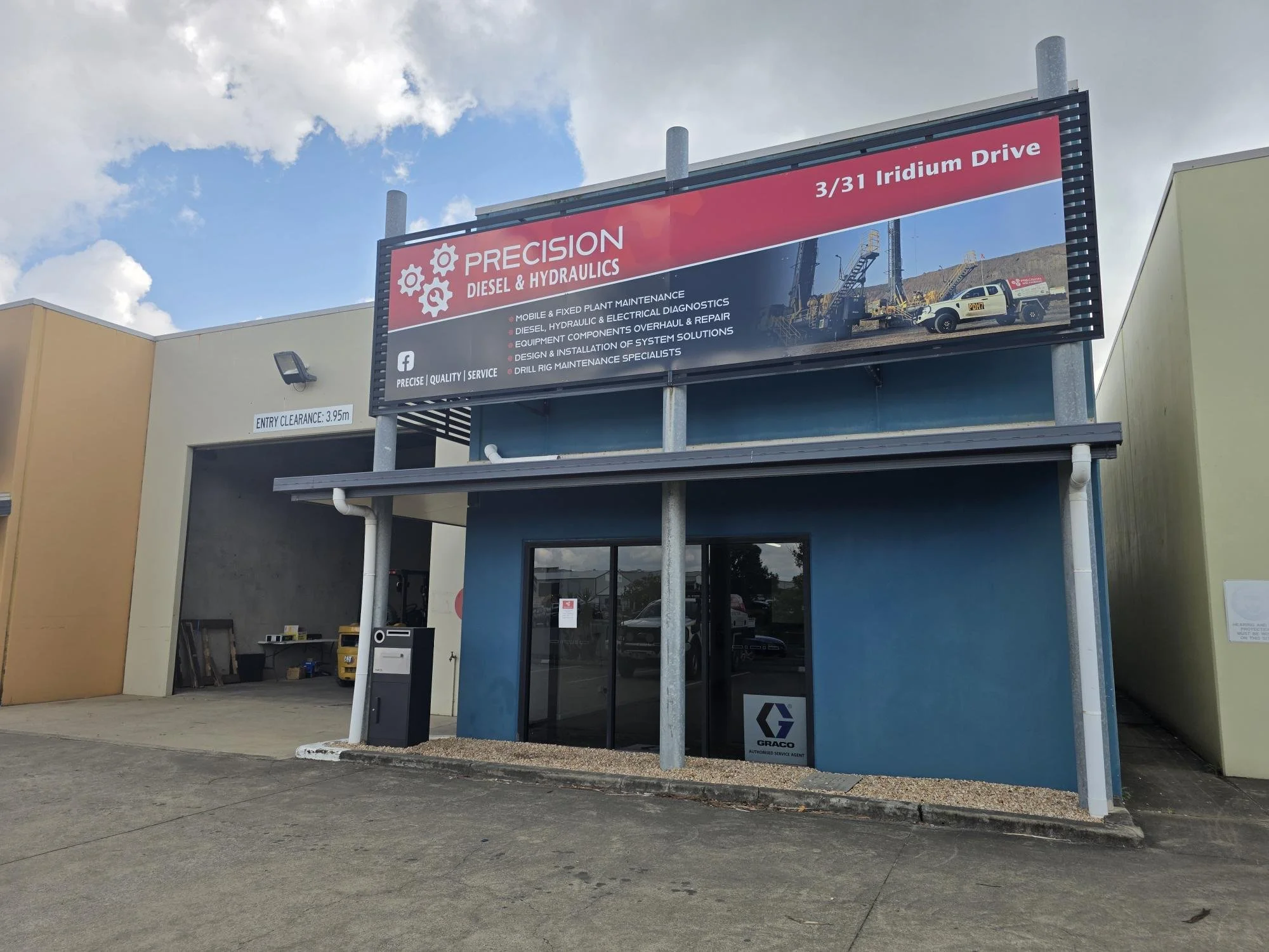 Commercial building with signage for Precision Diesel and Hydraulics, located at 3/31 Iridium Drive, featuring a large digital billboard advertising diesel and hydraulic services, with a glass door entrance and parking area in front.