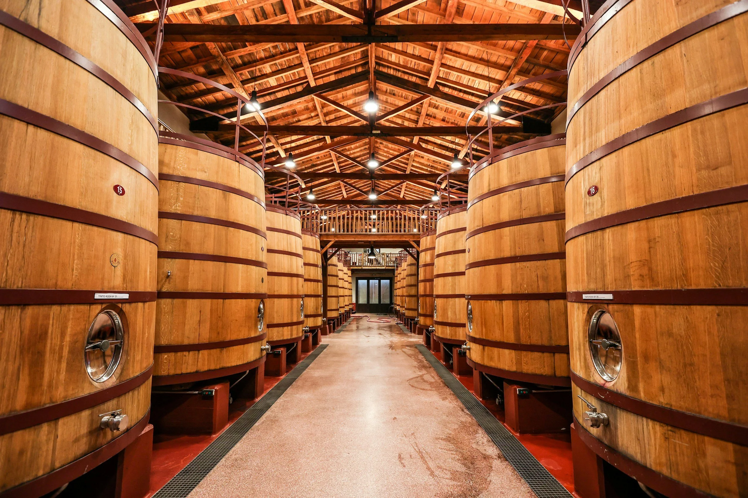 View of a wine cellar with large wooden fermentation tanks arranged on both sides of a central aisle, wooden ceiling, and a door at the end of the aisle.