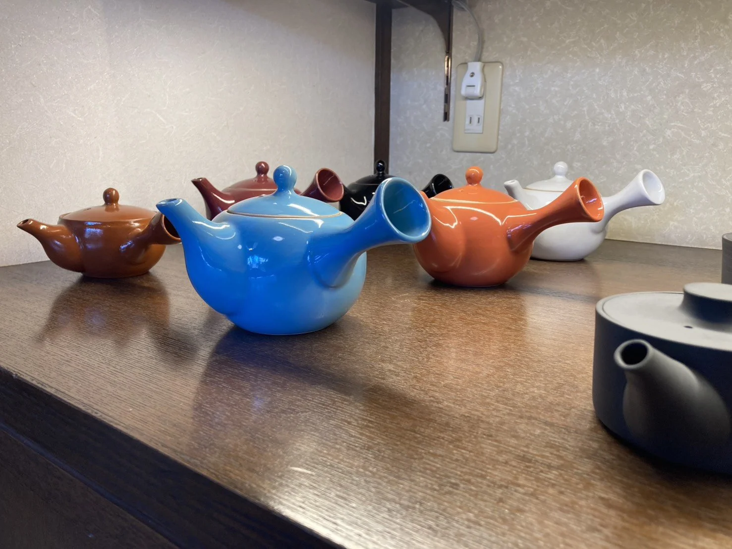 Colorful ceramic teapots in various colors including blue, white, black, brown, and orange are arranged on a wooden surface in front of a light-colored wall with a power outlet and an extension cord.