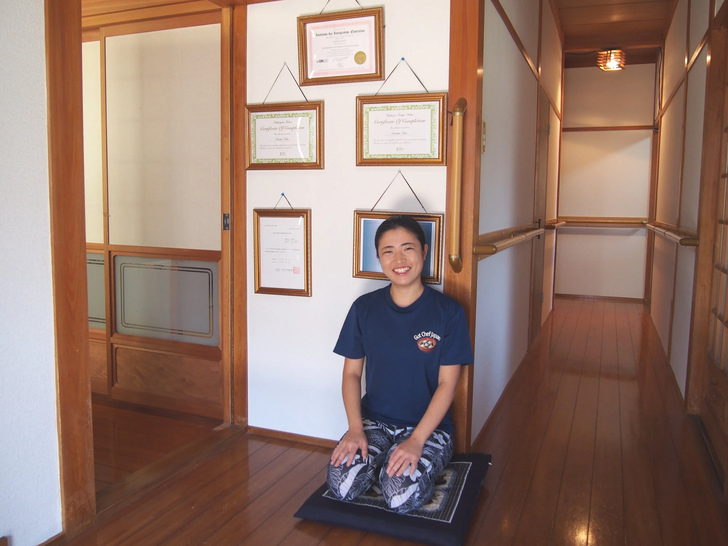 A woman kneeling on a mat in a hallway, smiling, with framed certificates and a portrait on the wall behind her.