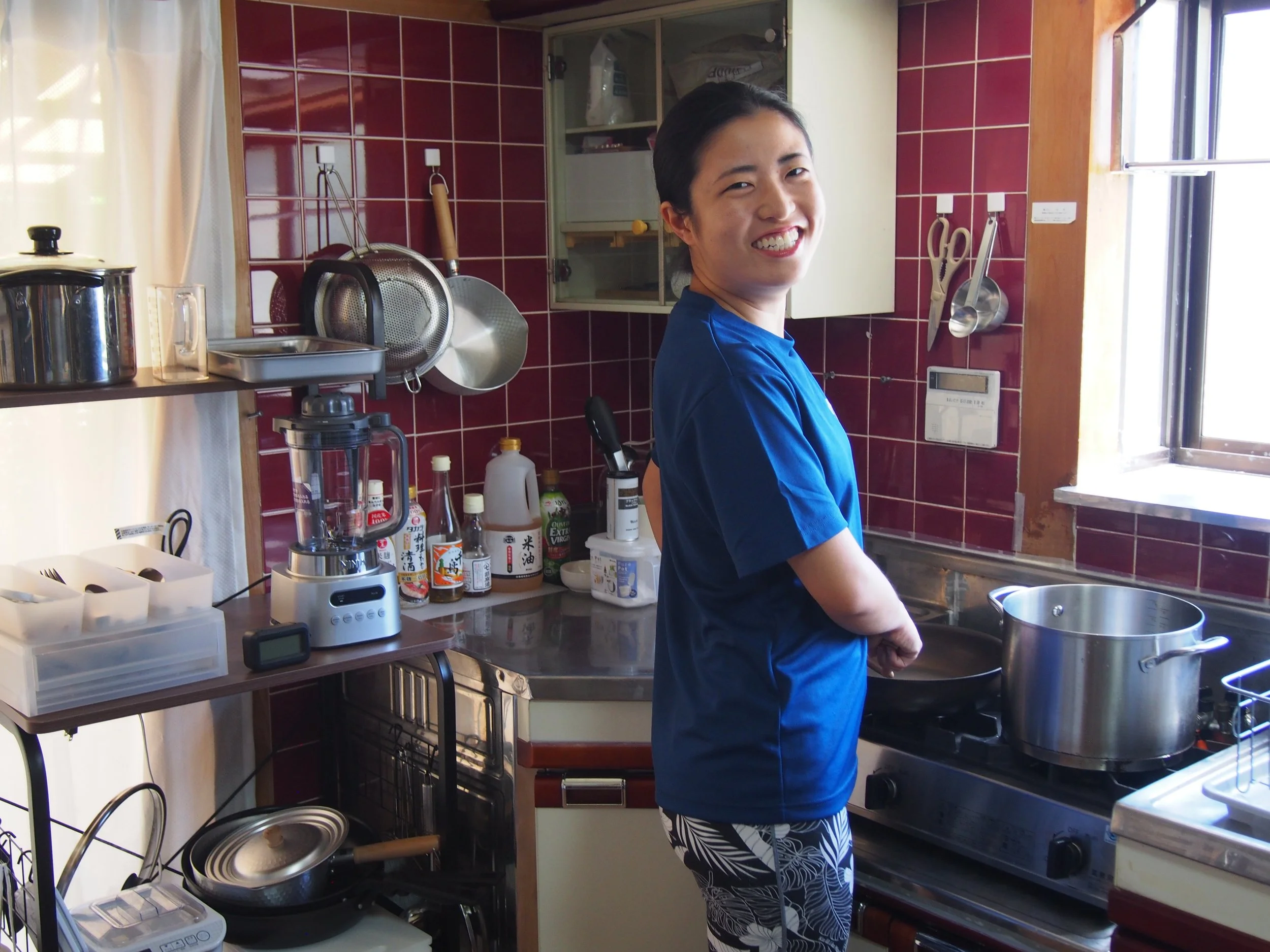 A smiling woman with black hair tied back, wearing a blue top and patterned black and white pants, appears to be working in a kitchen with red tiled walls, various kitchen utensils hanging on the wall, and kitchen appliances and bottles on the counter.