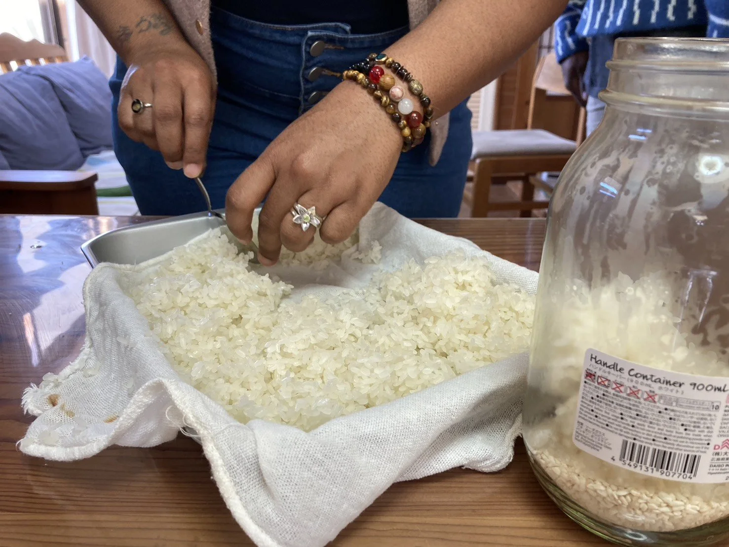 Person scooping rice from a cloth-lined container with a metal scoop, with a jar of rice nearby, on a wooden table.