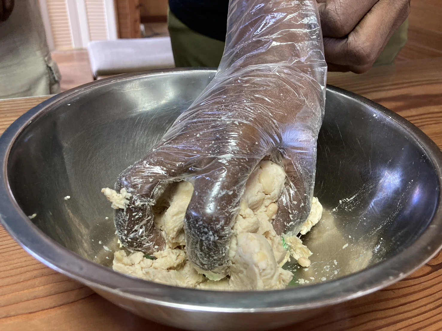 A person wearing a plastic glove mixing dough in a metal bowl on a wooden table.