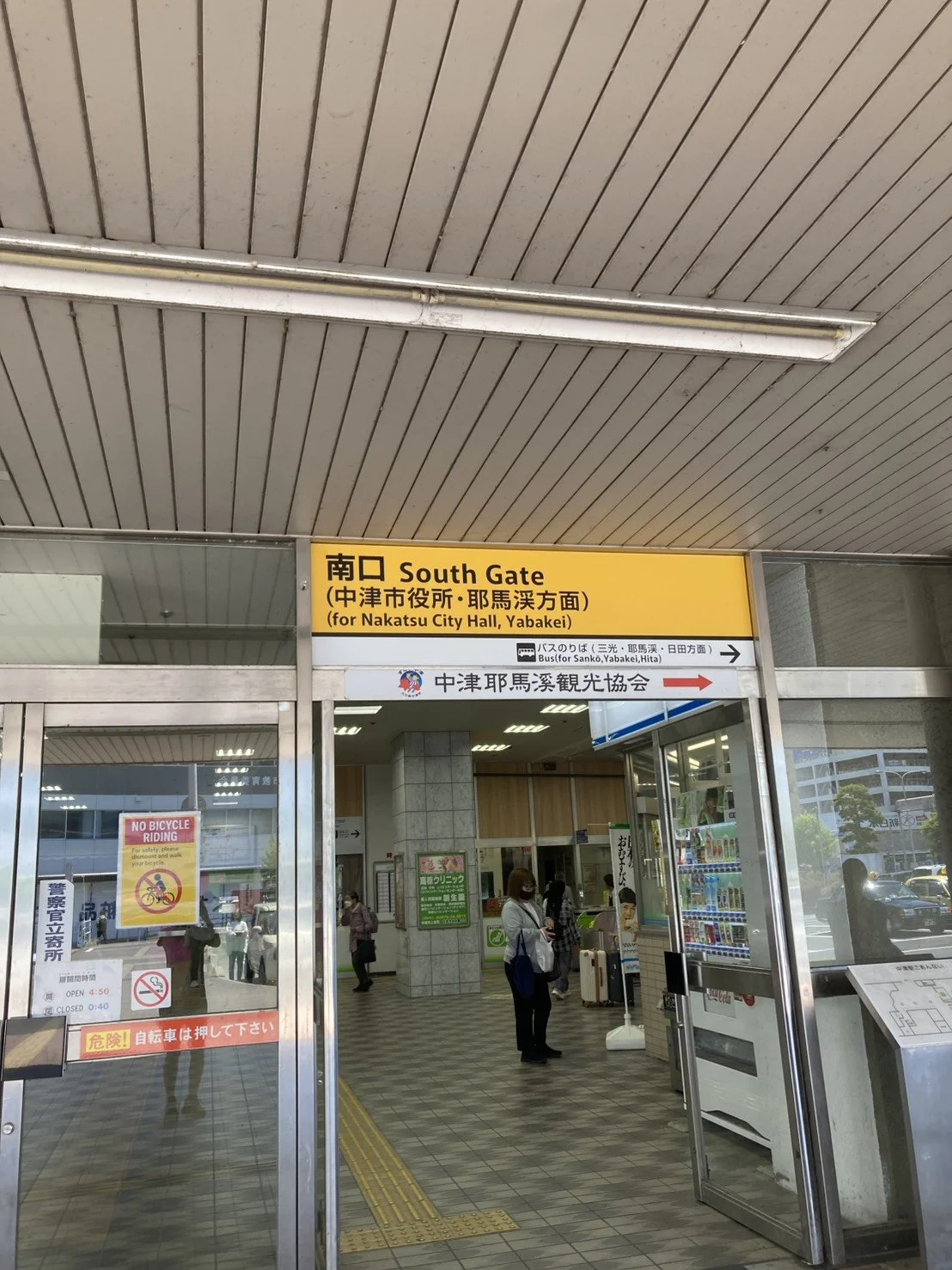 Entrance to the South Gate of a train station with signs indicating directions for Nakatsu City Hall, Yabakei, and bus routes. Several people are seen inside and outside the station, some wearing masks.