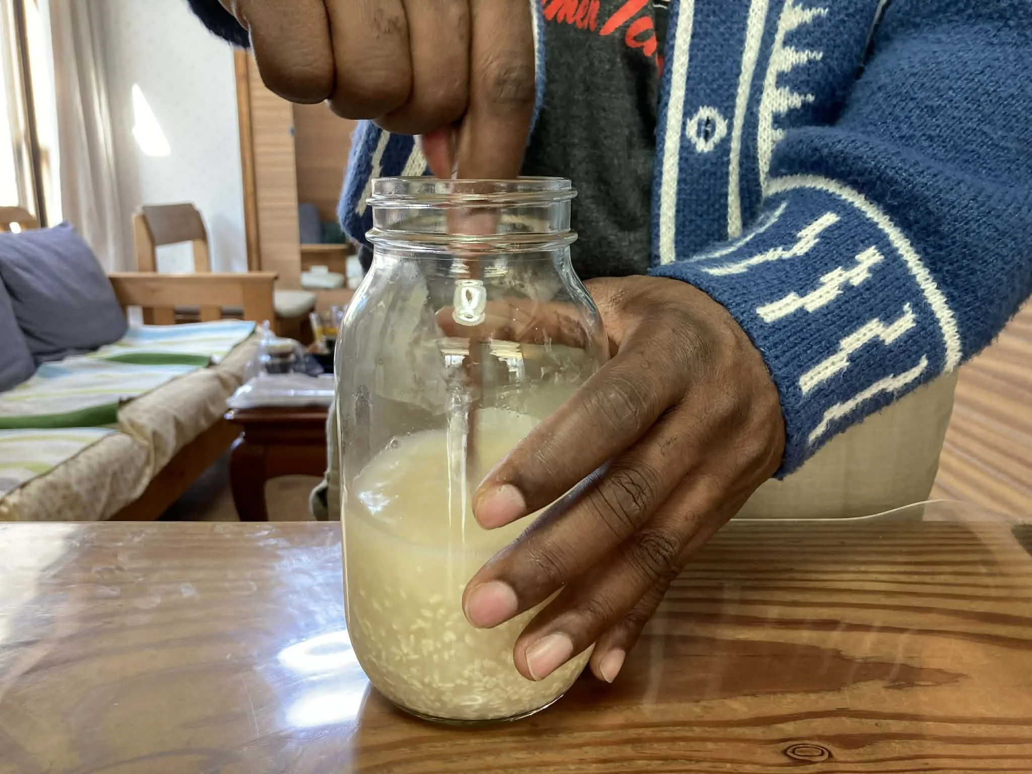 A person pouring liquid from a spoon into a glass jar with a lemon slice inside, on a wooden table in a cozy room.
