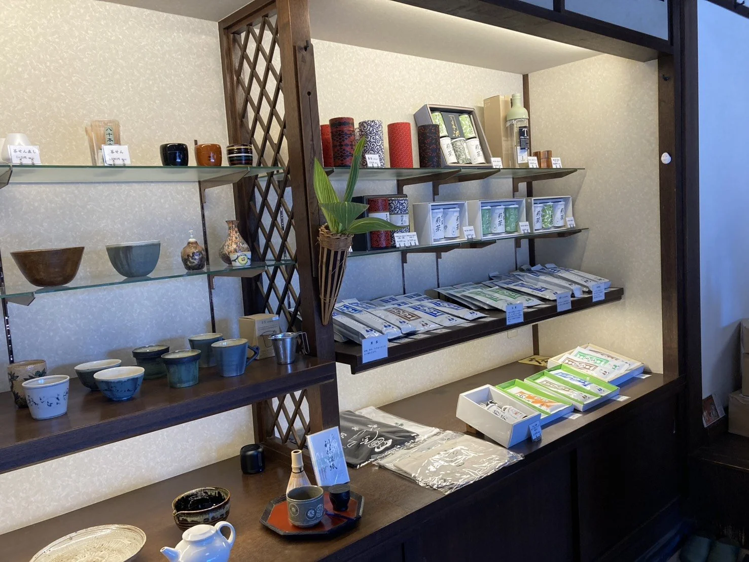 Display shelf with various ceramic bowls, cups, and vases, and packaged tea products in a store with a wooden lattice divider.