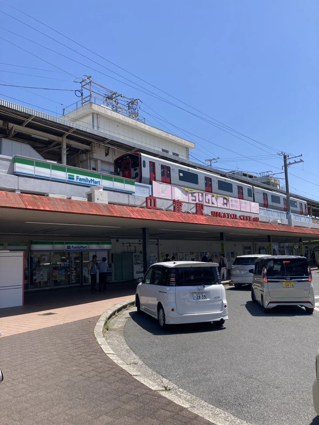 A train on an elevated track above a bus station with parked cars and pedestrians, with clear blue sky in the background.