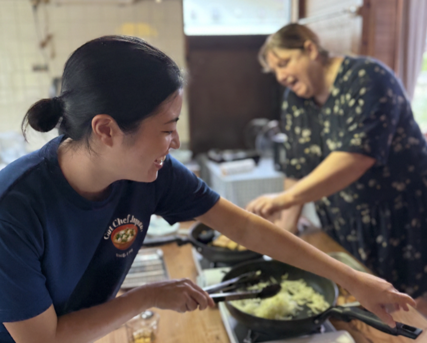 Two women happily cooking together in a kitchen. One woman is stirring a pot of rice, while the other is chopping or preparing food in the background.