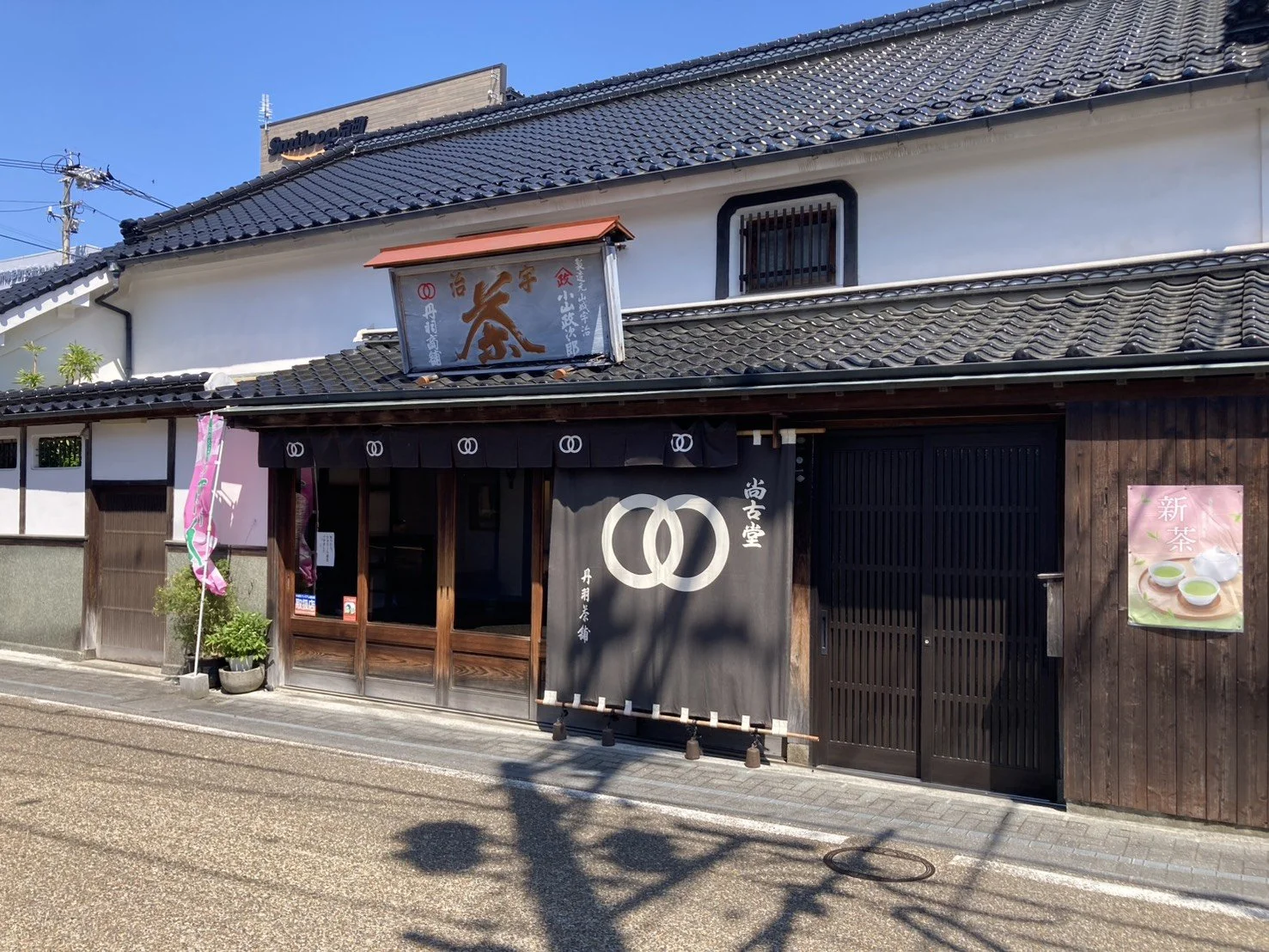 Traditional Japanese building with dark wooden exterior, black tiled roof, and white curtains featuring white intertwined loops, possibly a tea shop, with colorful banners and a poster advertising green tea on the right side.