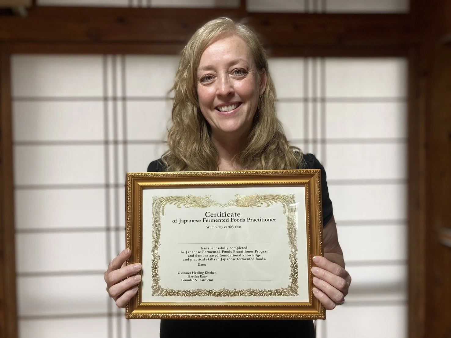 A woman with long blonde hair smiling and holding a framed certificate of completion for a Japanese fermented foods practitioner program, standing in front of a traditional Japanese shoji screen.
