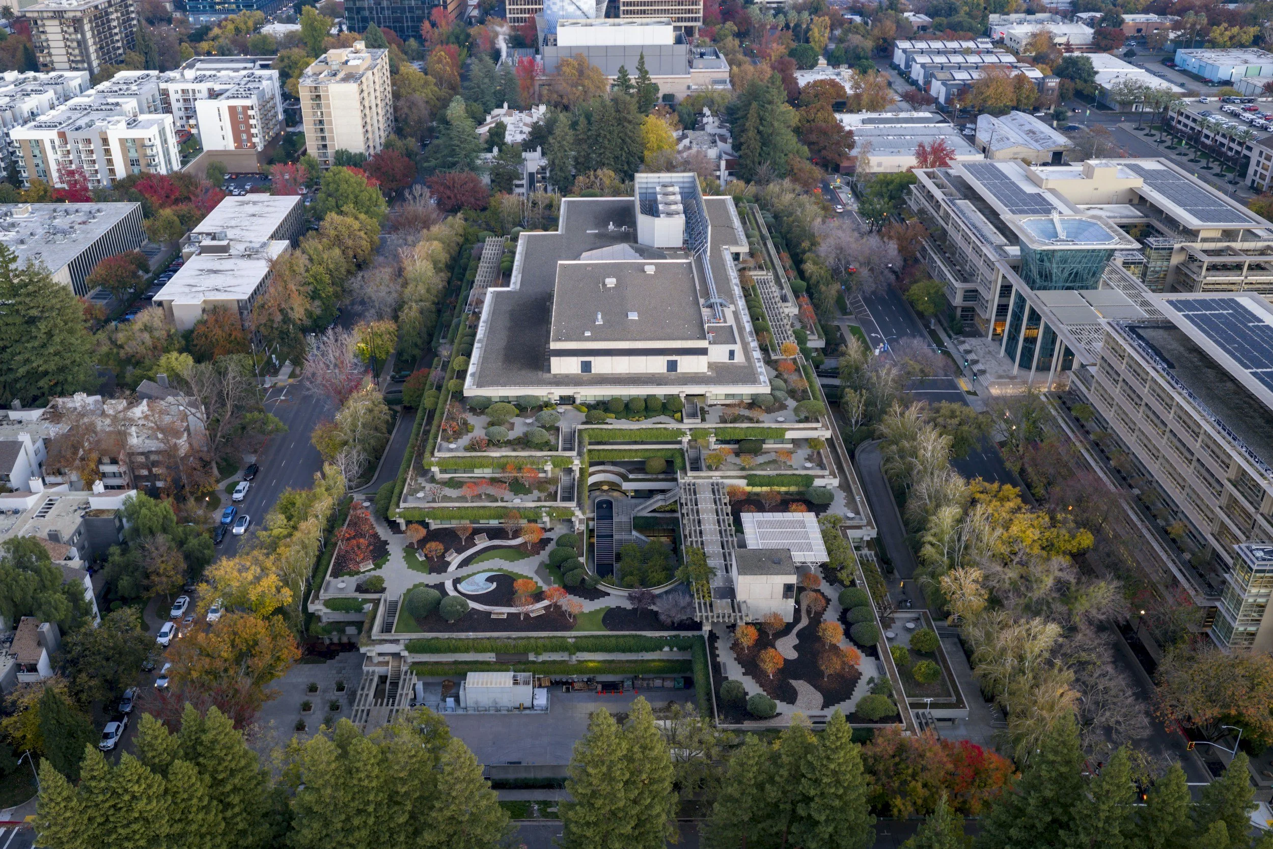 Aerial view of an urban area featuring a large multistory building with a landscaped rooftop garden, surrounded by trees, roads, and other commercial buildings.