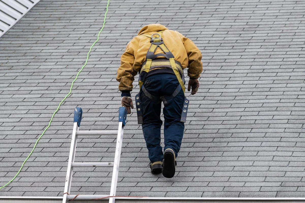 Construction worker wearing safety harness climbing a sloped gray shingled roof, with a ladder nearby and a green safety line attached.