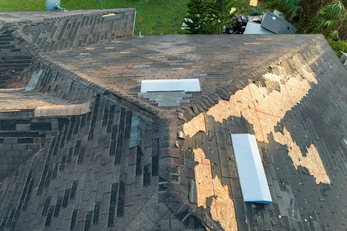 Aerial view of a house roof with damaged shingles revealing underlayment beneath, and two repairs with new white vent covers.
