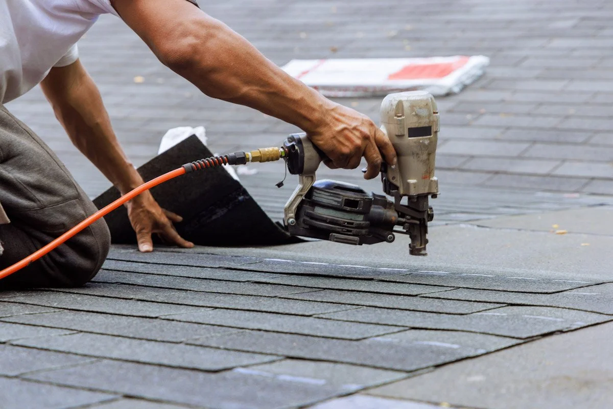 Worker installing asphalt shingles on a roof using a pneumatic nail gun.