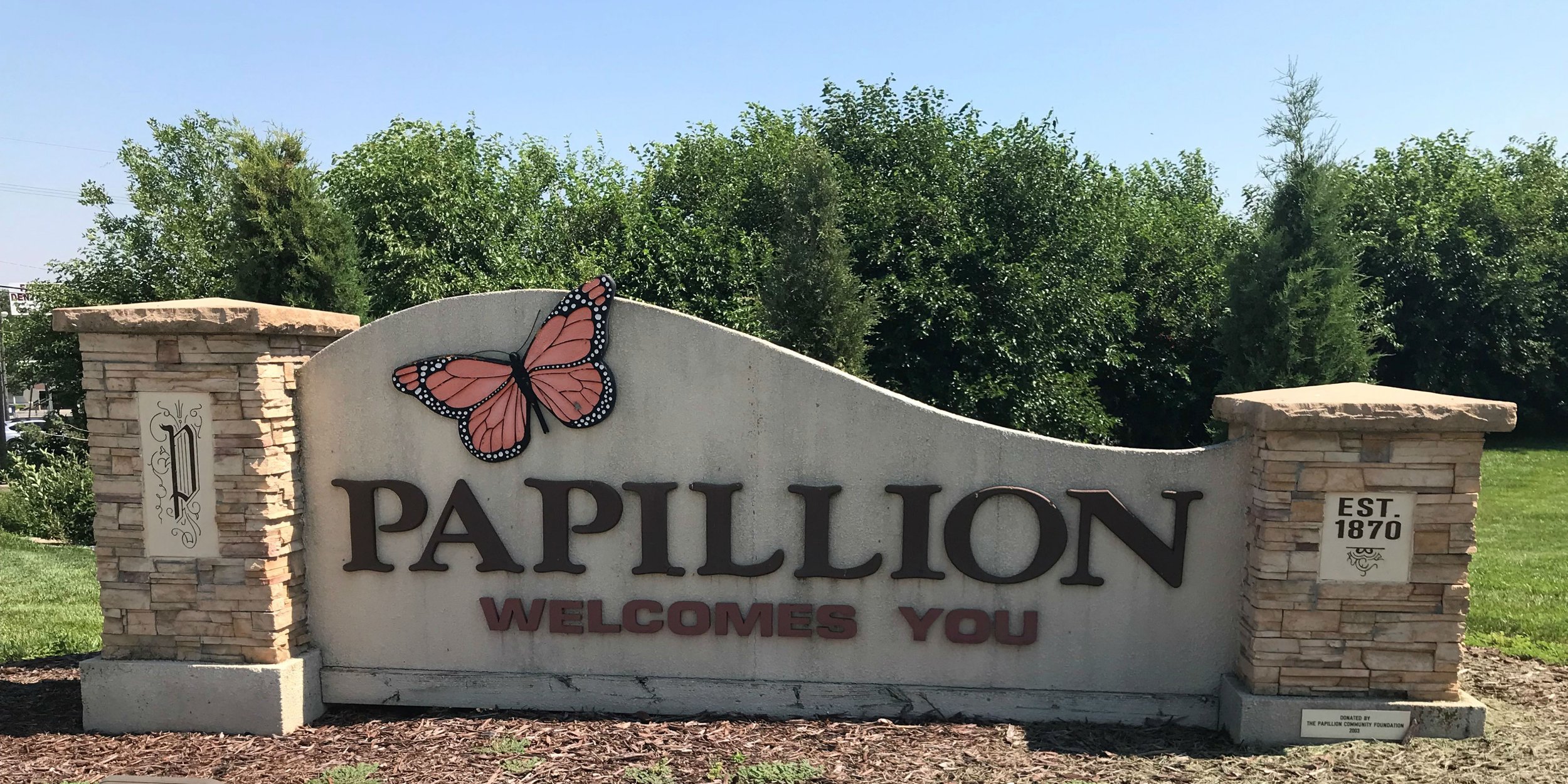 A welcoming sign for Papillion, featuring a pink and black monarch butterfly, with stone pillars on each side, set against green trees and a clear blue sky.