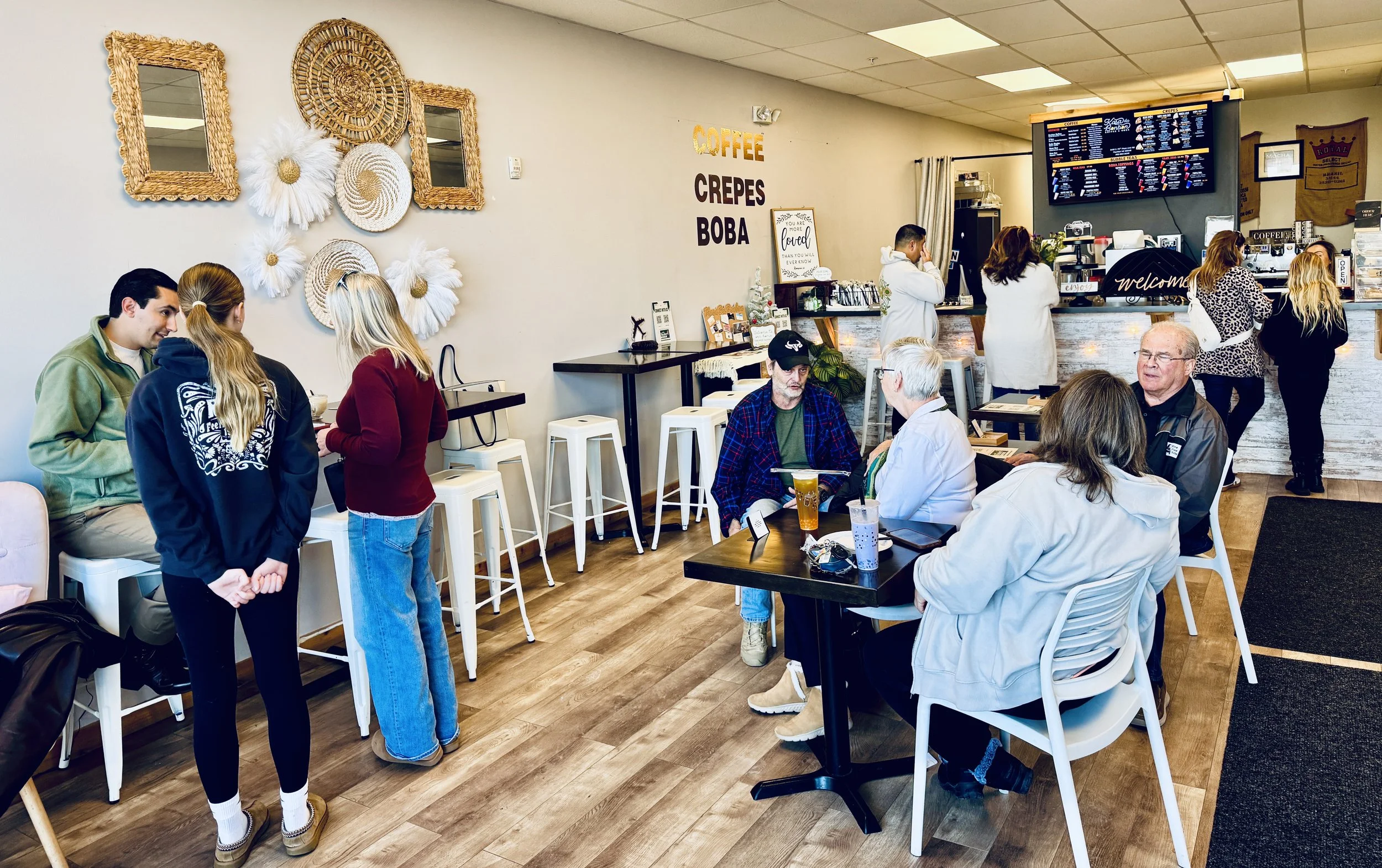 A busy coffee shop with people standing and sitting, a menu on a digital screen, decorated wall art, and a counter with coffee machines.