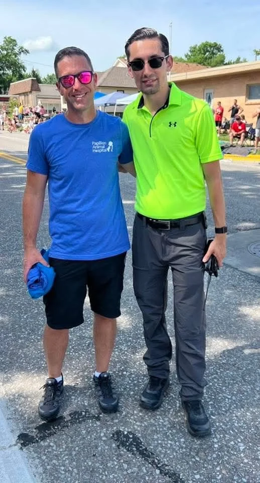 Two men standing together outdoors during a community event. One man wears a blue t-shirt with a logo and black shorts, holding a blue garment, while the other wears a bright yellow-green polo shirt and dark gray pants. Both are wearing sunglasses and smiling.