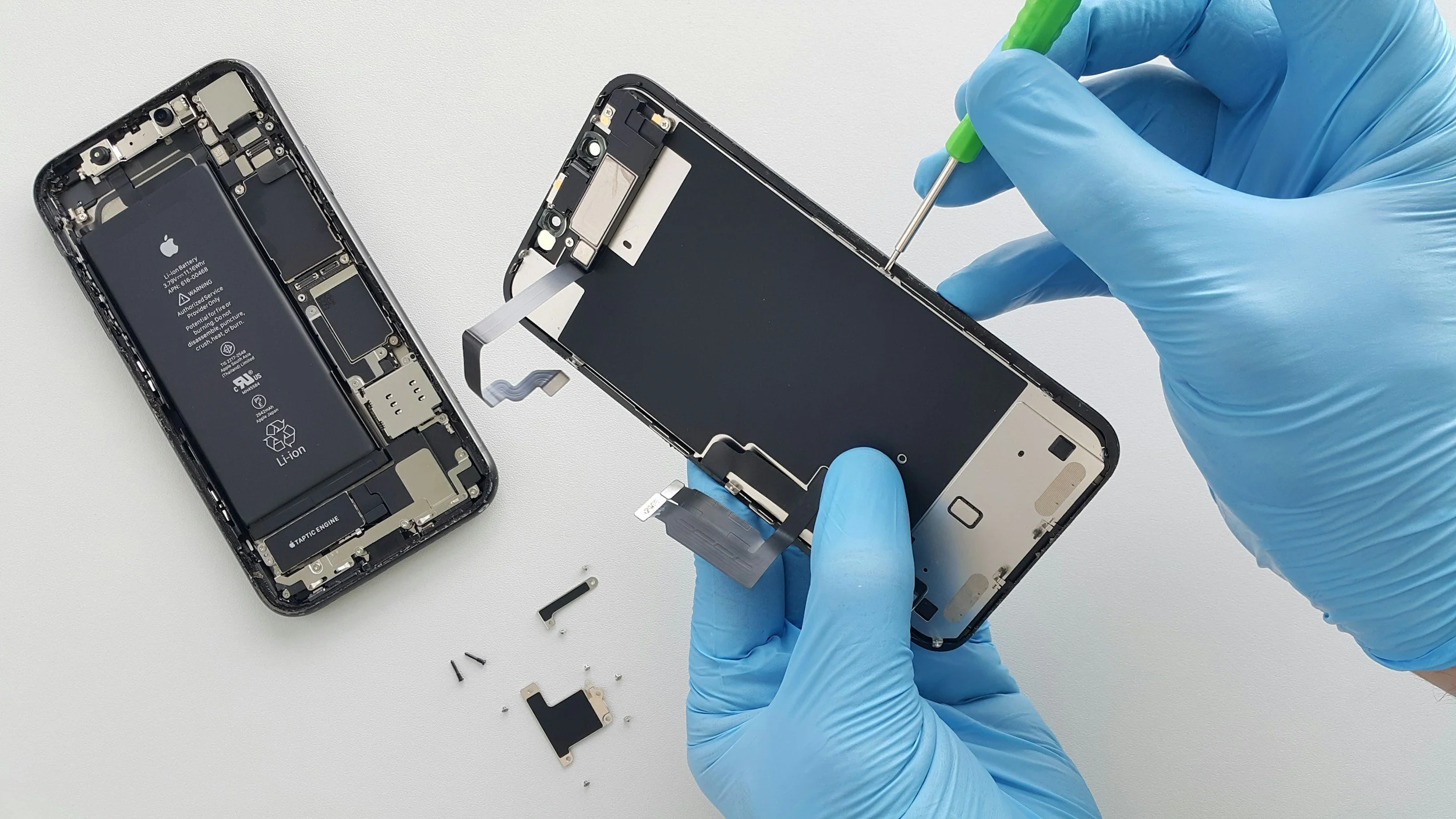 A person wearing blue gloves is repairing a disassembled smartphone with parts and tools on a white surface.