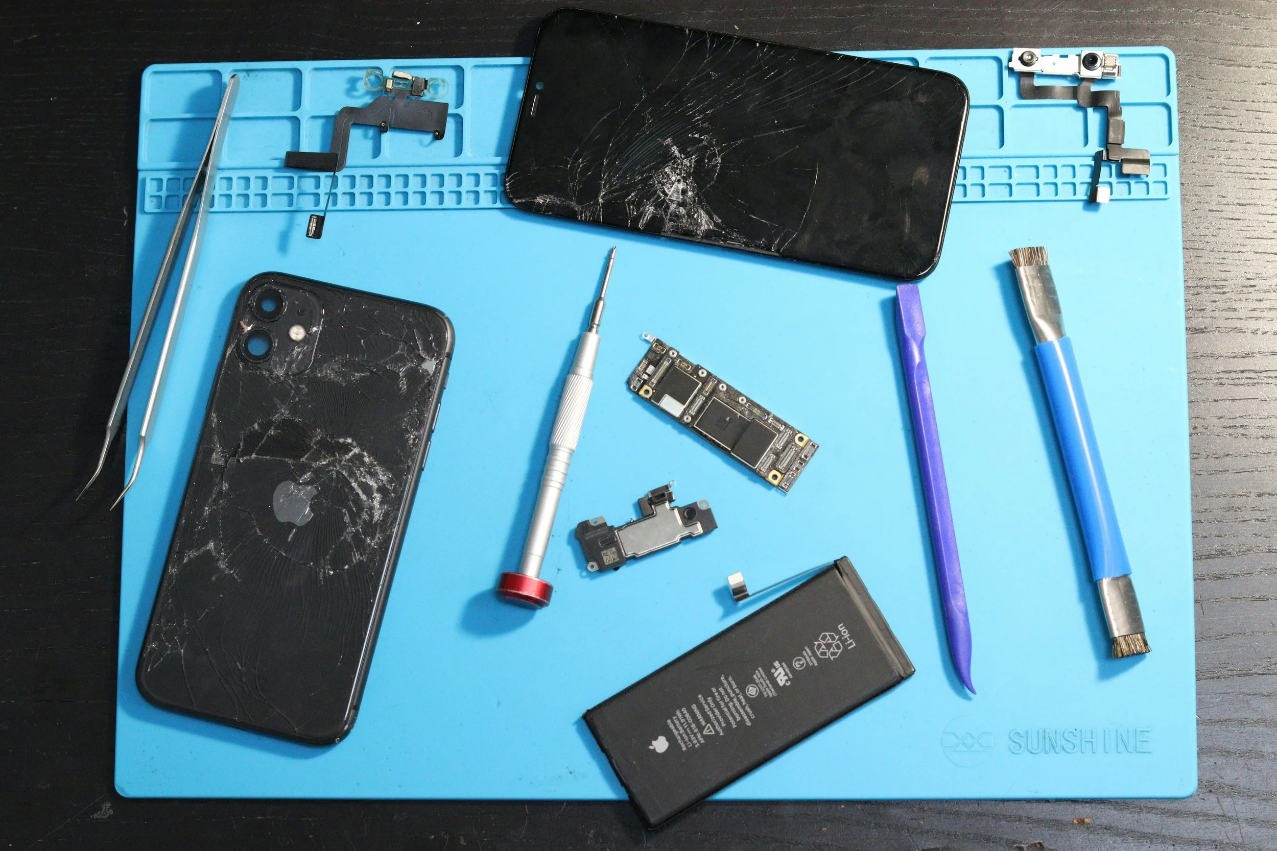 Disassembled black iPhone with cracked screen on a blue work mat surrounded by repair tools, including screwdrivers, tweezers, and a replacement battery.