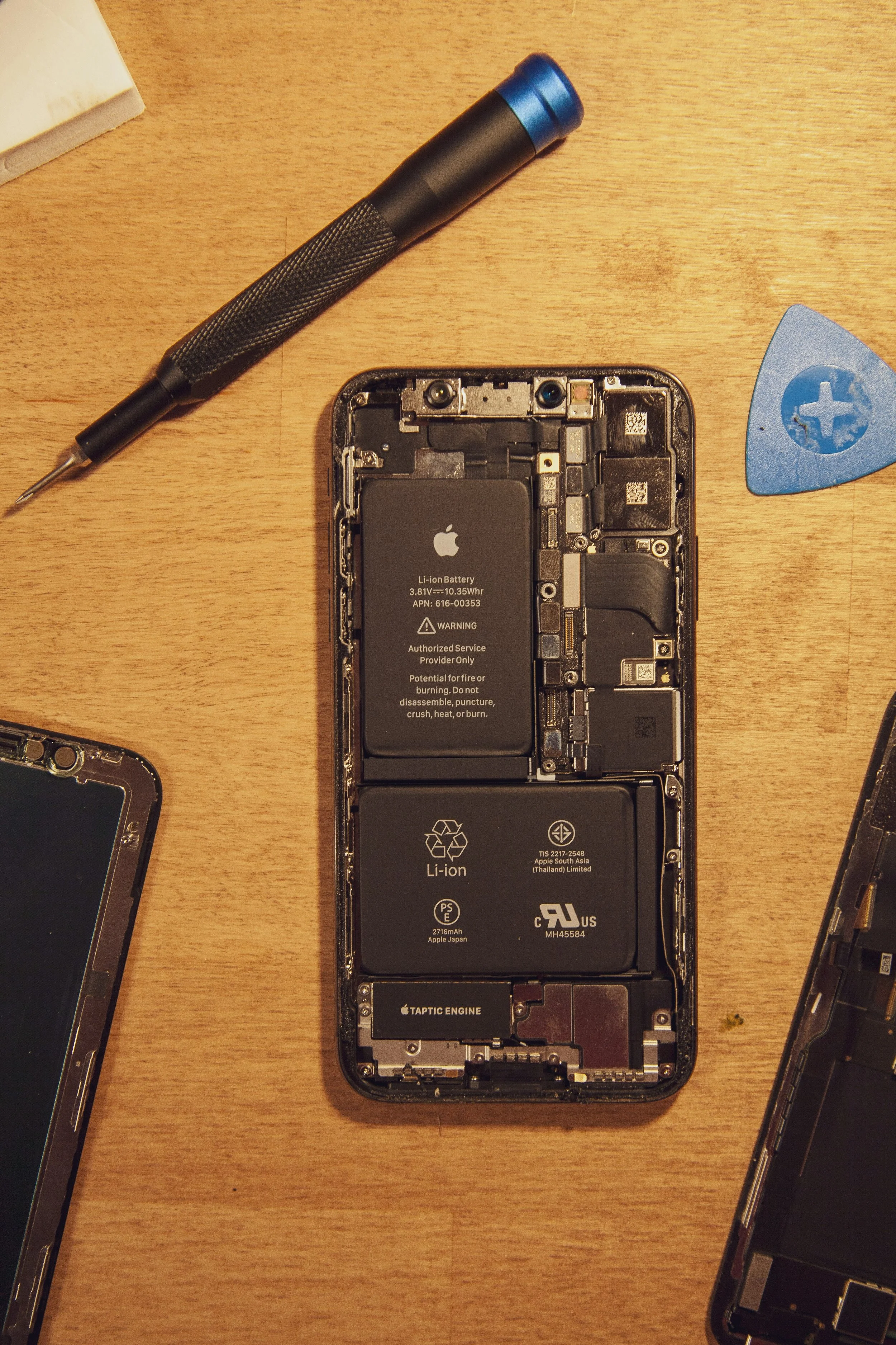 Inside view of a disassembled iPhone showing the battery, circuitry, and components, with a toolkit and a guitar pick nearby on a wooden surface.
