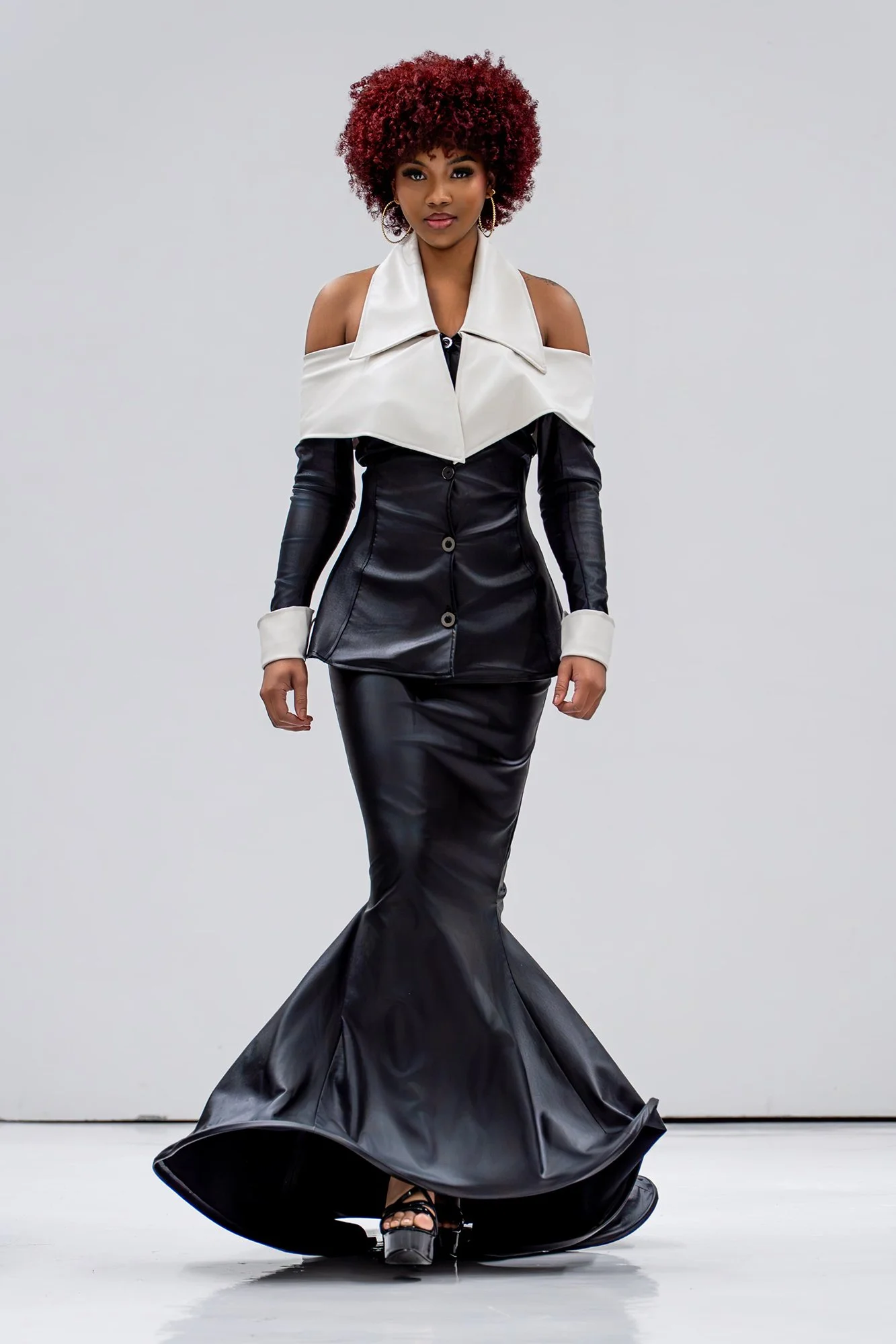 Model walking on runway in a black leather gown with a mermaid silhouette, paired with a white off-shoulder jacket with large lapels, and high heels.