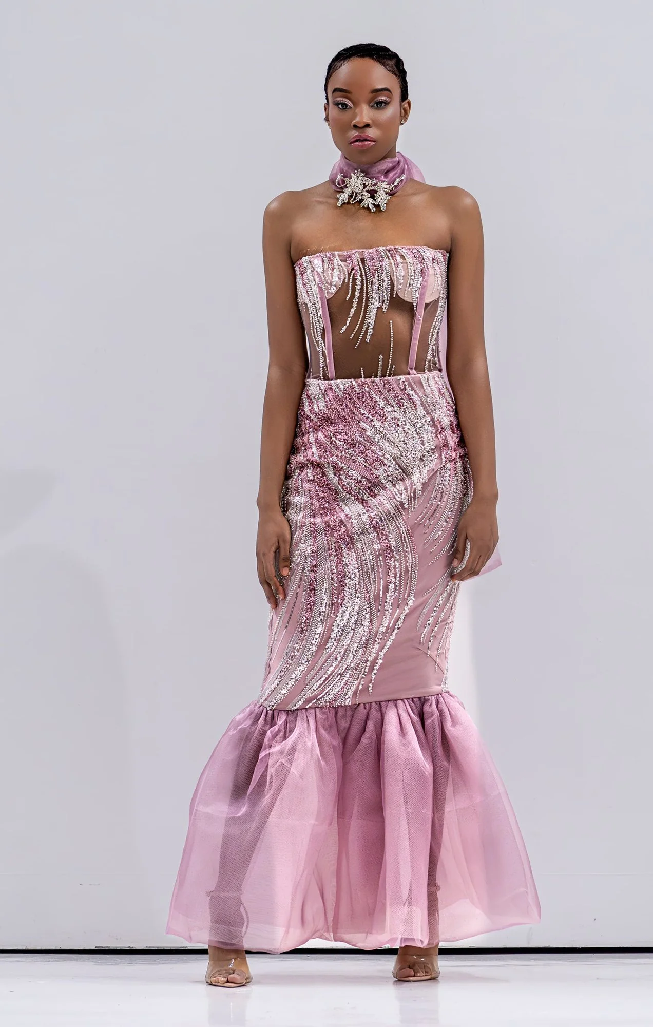 A woman wearing a strapless pink sequined dress with a sheer midsection and a ruffled tulle hem, standing against a plain background.