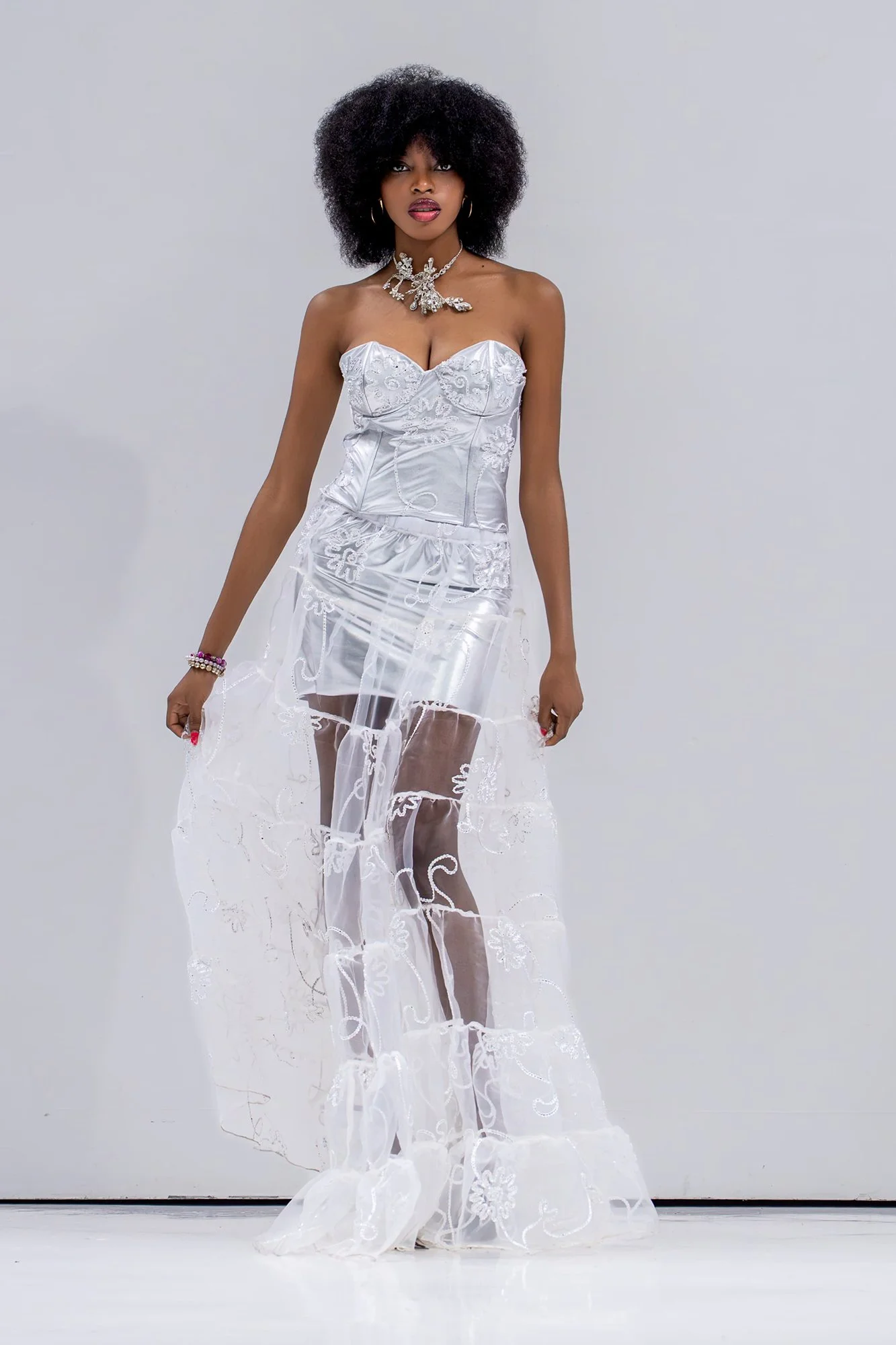 A woman with dark skin and curly hair is wearing a white, strapless dress with intricate embroidery. The dress has a fitted bodice and a sheer, embroidered overlay that extends to the floor. She is accessorized with large hoop earrings, a statement n