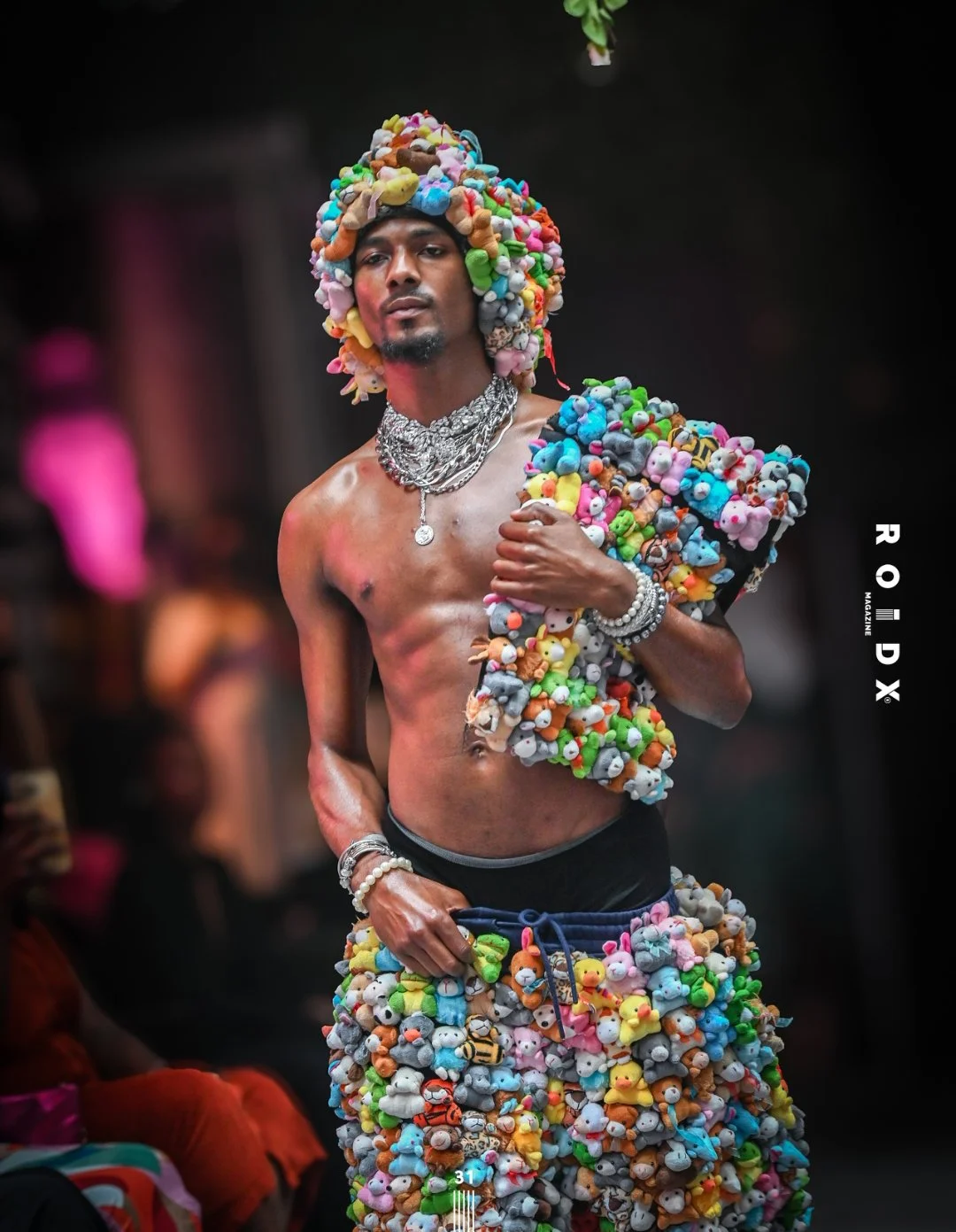 Man with dark skin and a goatee, shirtless, wearing an elaborate hat and outfit covered in colorful plush teddy bears, holding his left arm across his chest, with jewelry including necklaces and bracelets, walking on a runway.