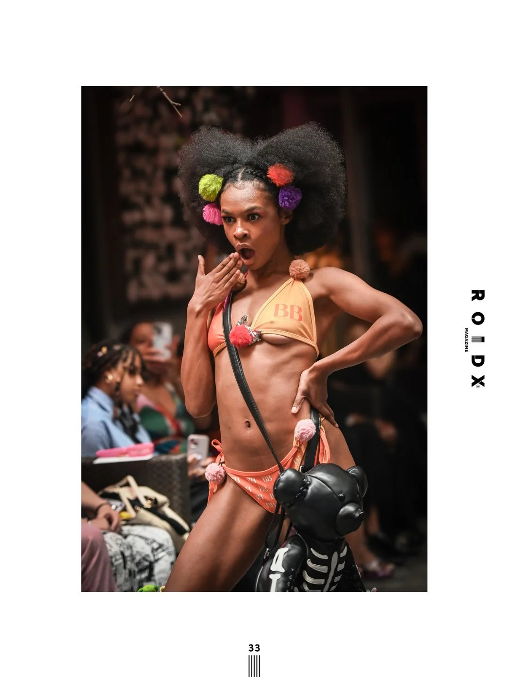 A woman with a surprised expression, wearing colorful pom-pom hair accessories, a bikini with BB branding, and carrying a black bear-shaped bag, walking on a runway at a fashion show.