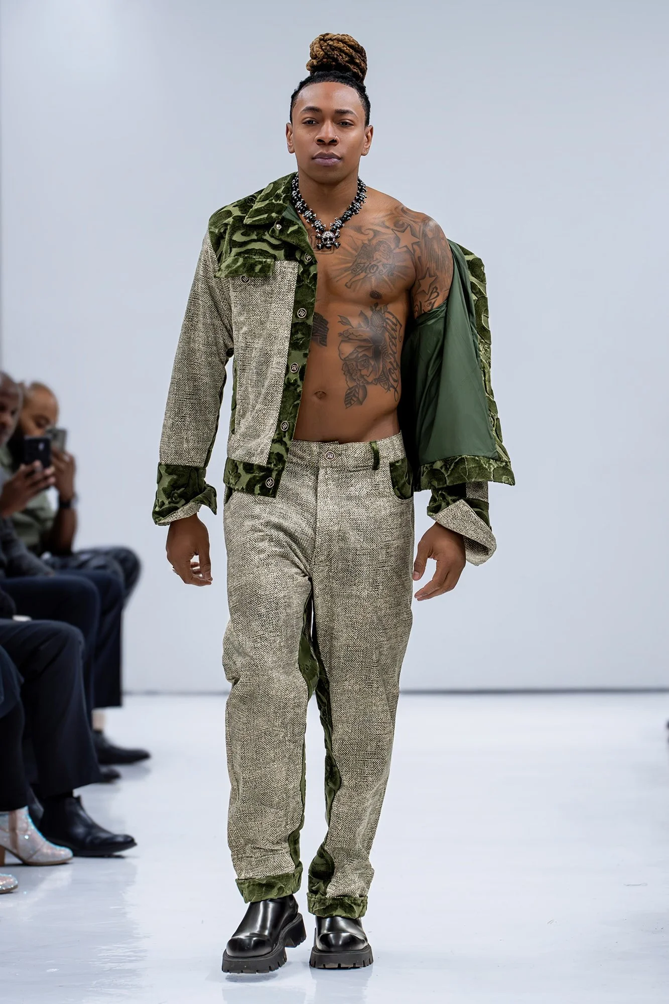 Male model walking on runway, wearing patterned jacket, green and beige pants, and black boots, with an audience watching in the background.