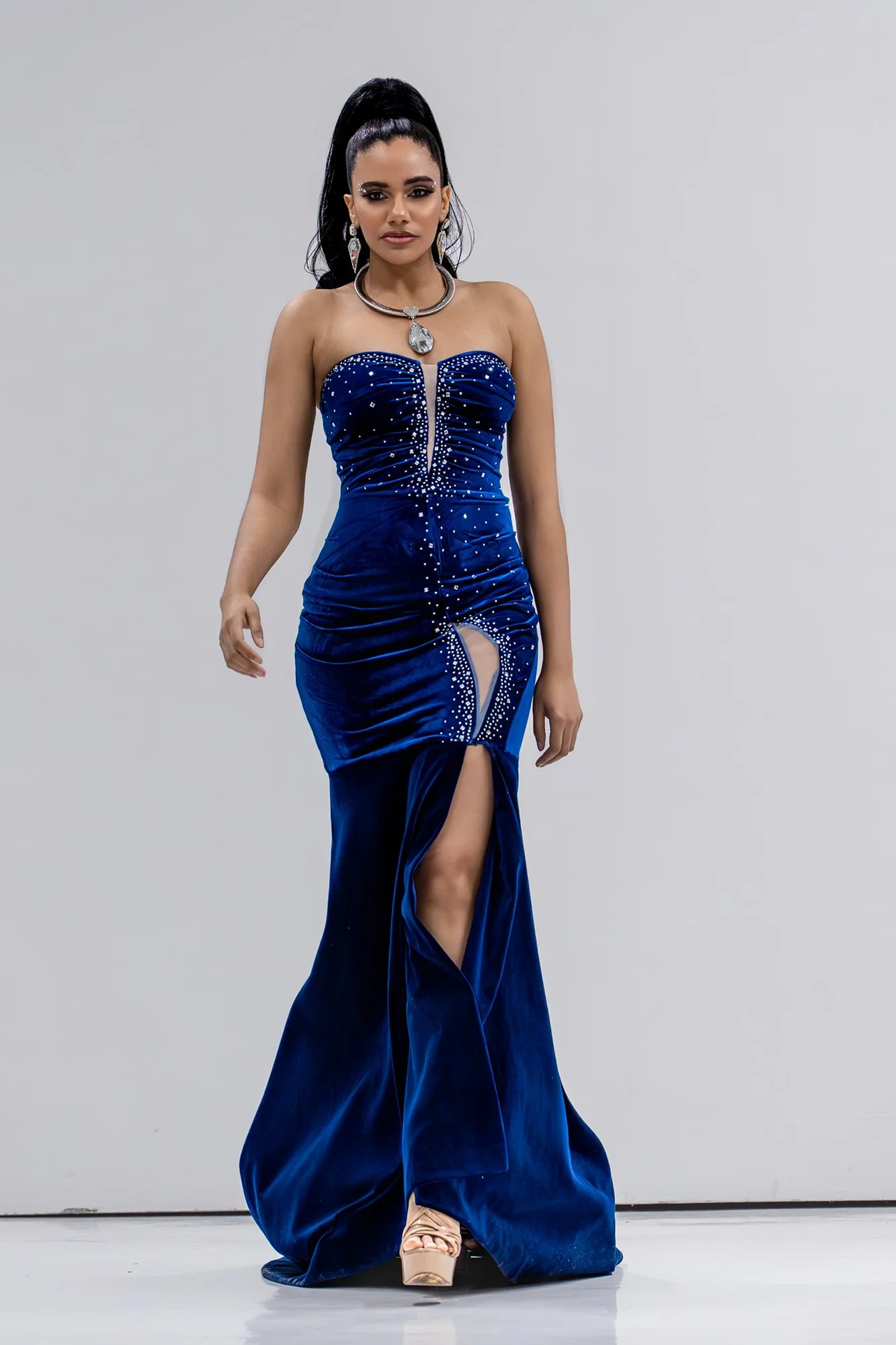 A woman in a strapless, blue, embellished evening gown with a slit revealing a leg, standing against a plain background.