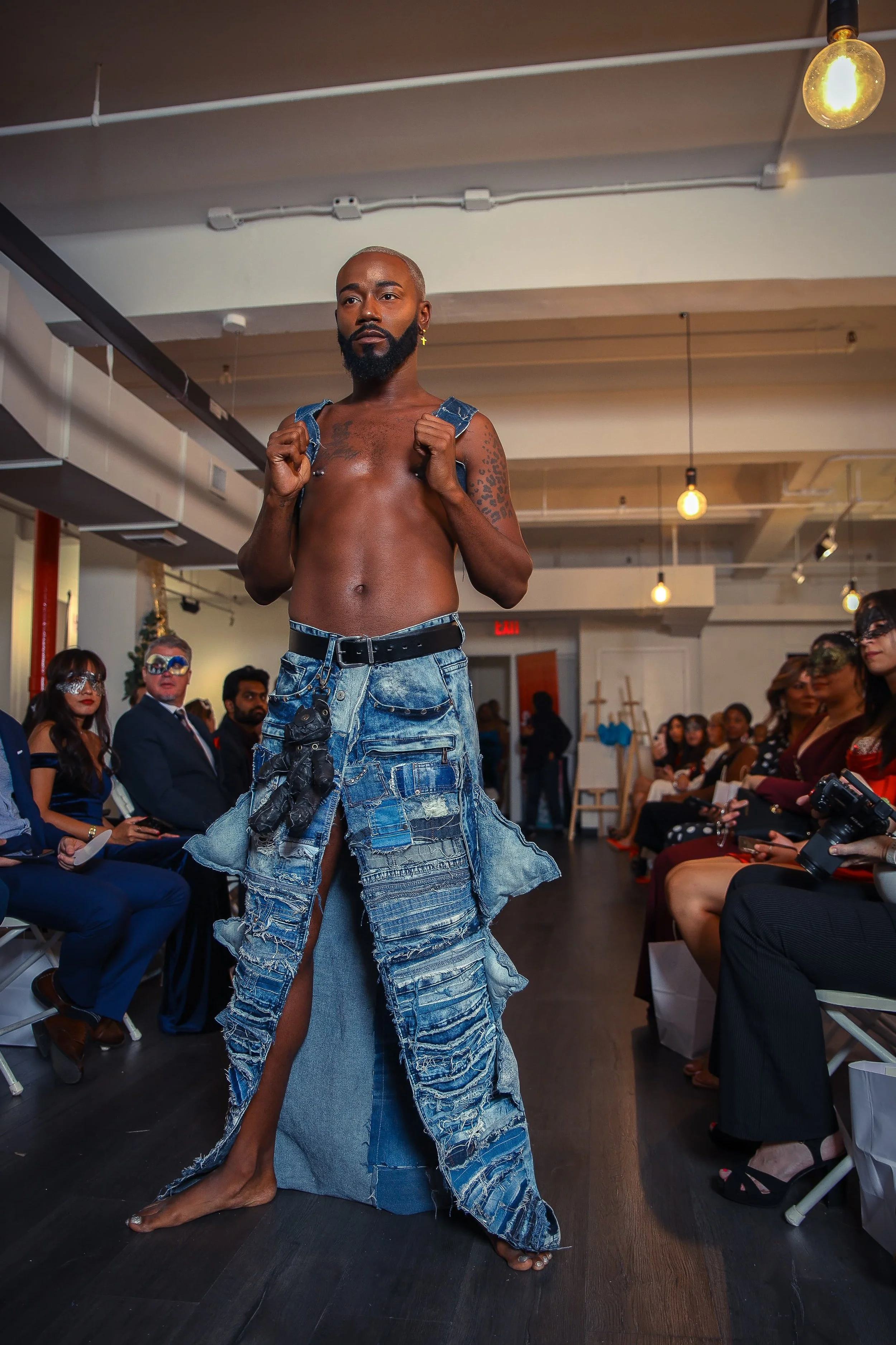 Man walking on runway at a fashion show, wearing ripped jeans with a long slit, no shirt, carrying a backpack, with an audience wearing masks seated on either side.