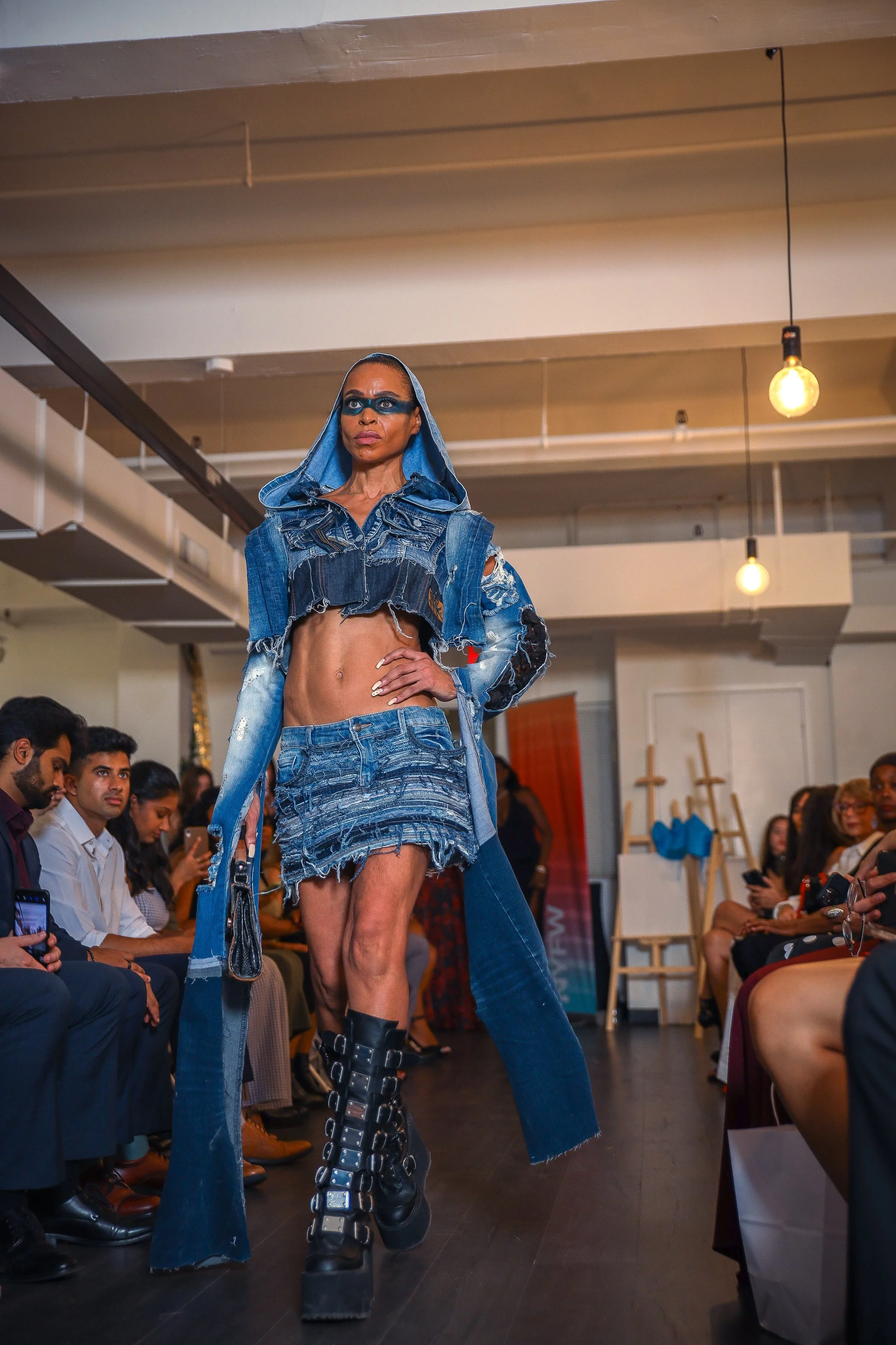 A female model walks down the runway wearing a distressed denim outfit, including a hooded jacket and a short skirt, paired with black chunky knee-high boots with buckles. The model has makeup with a bold black mask around her eyes and is striking a 